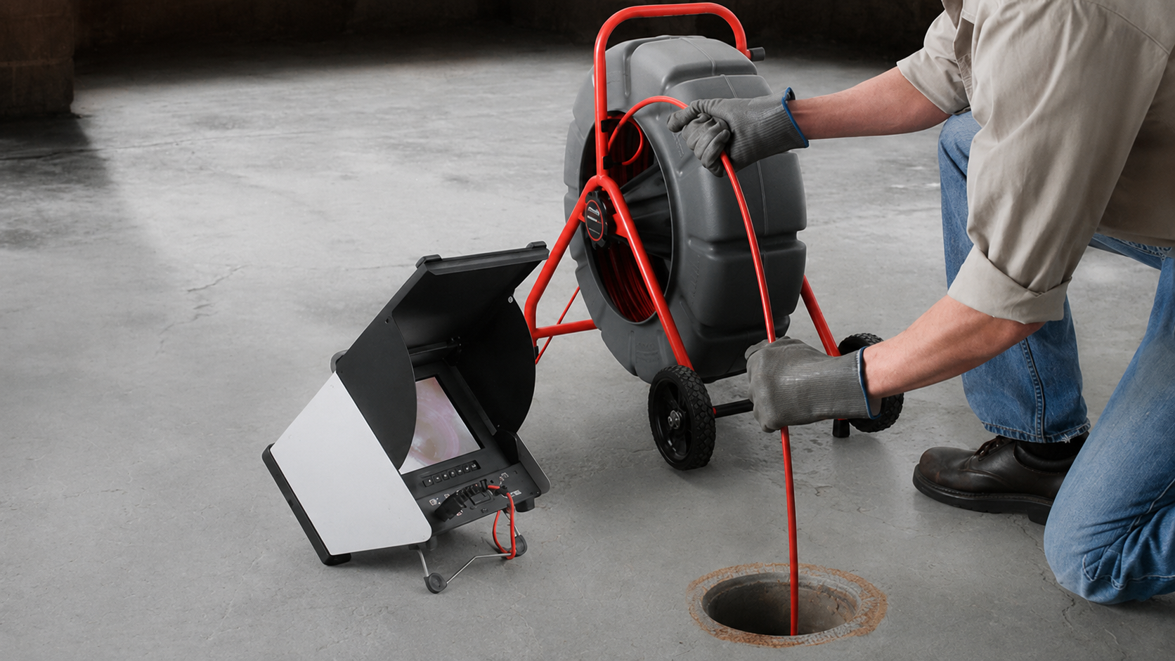 Worker lowers a wheeled machine into a round manhole with a red cable attached.
