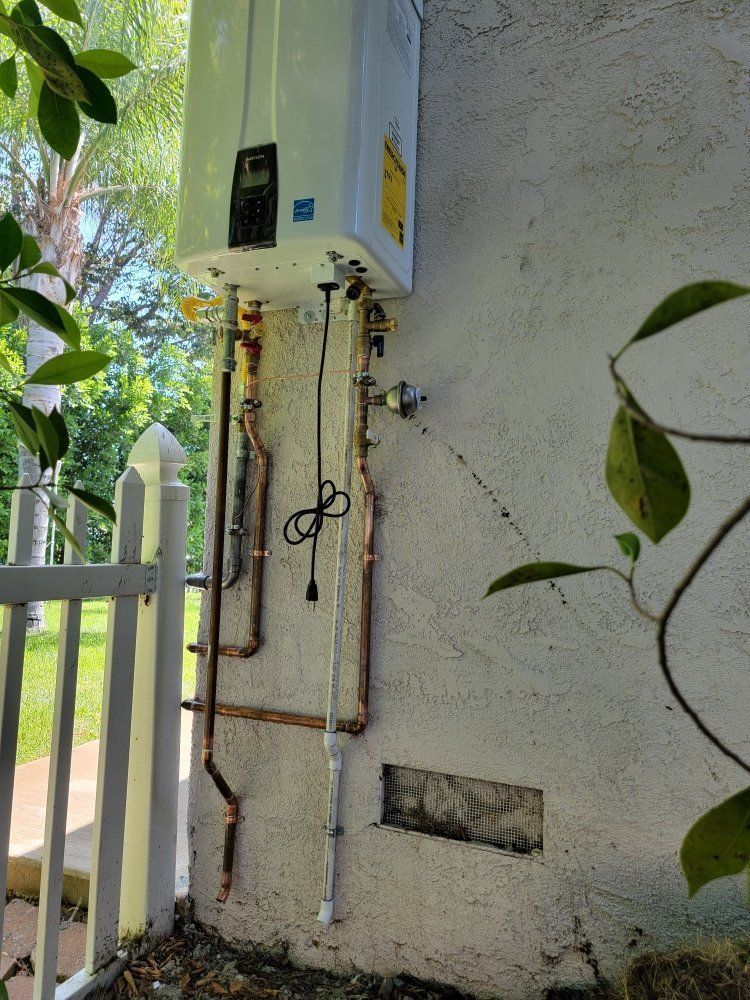 Outdoor utility meter box and exposed pipes mounted on a stucco wall beside a white fence.