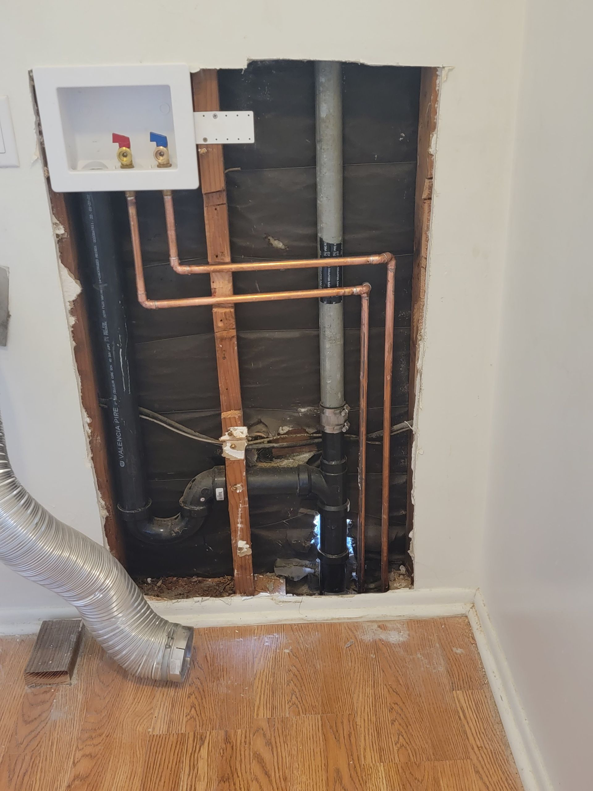 Exposed wall plumbing with copper pipes, vent pipe, and a flexible duct opening above hardwood floor