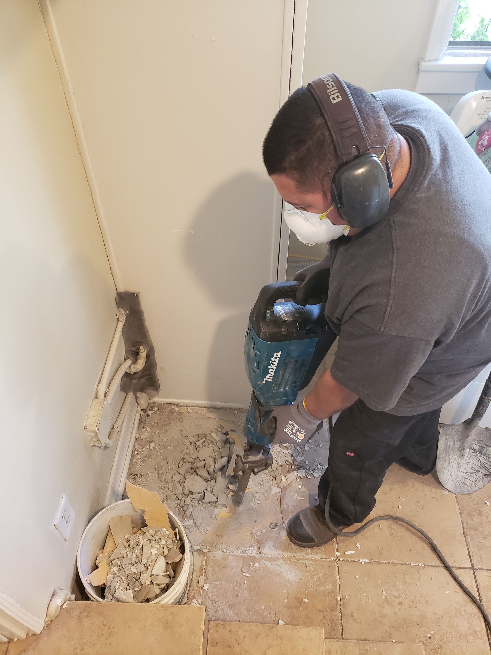 Worker using a power drill near a damaged wall and floor in a room with debris.