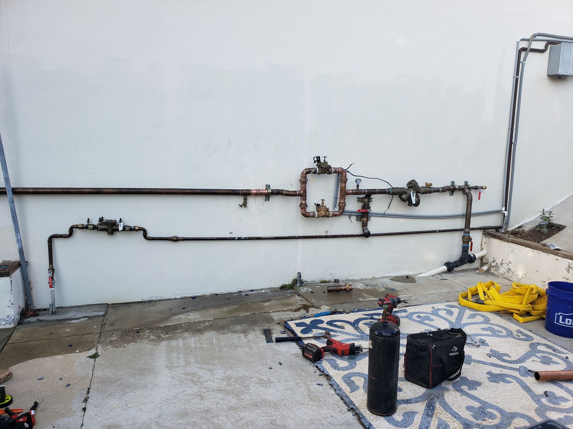Outdoor utility pipes along a white wall with tools and materials on the ground.
