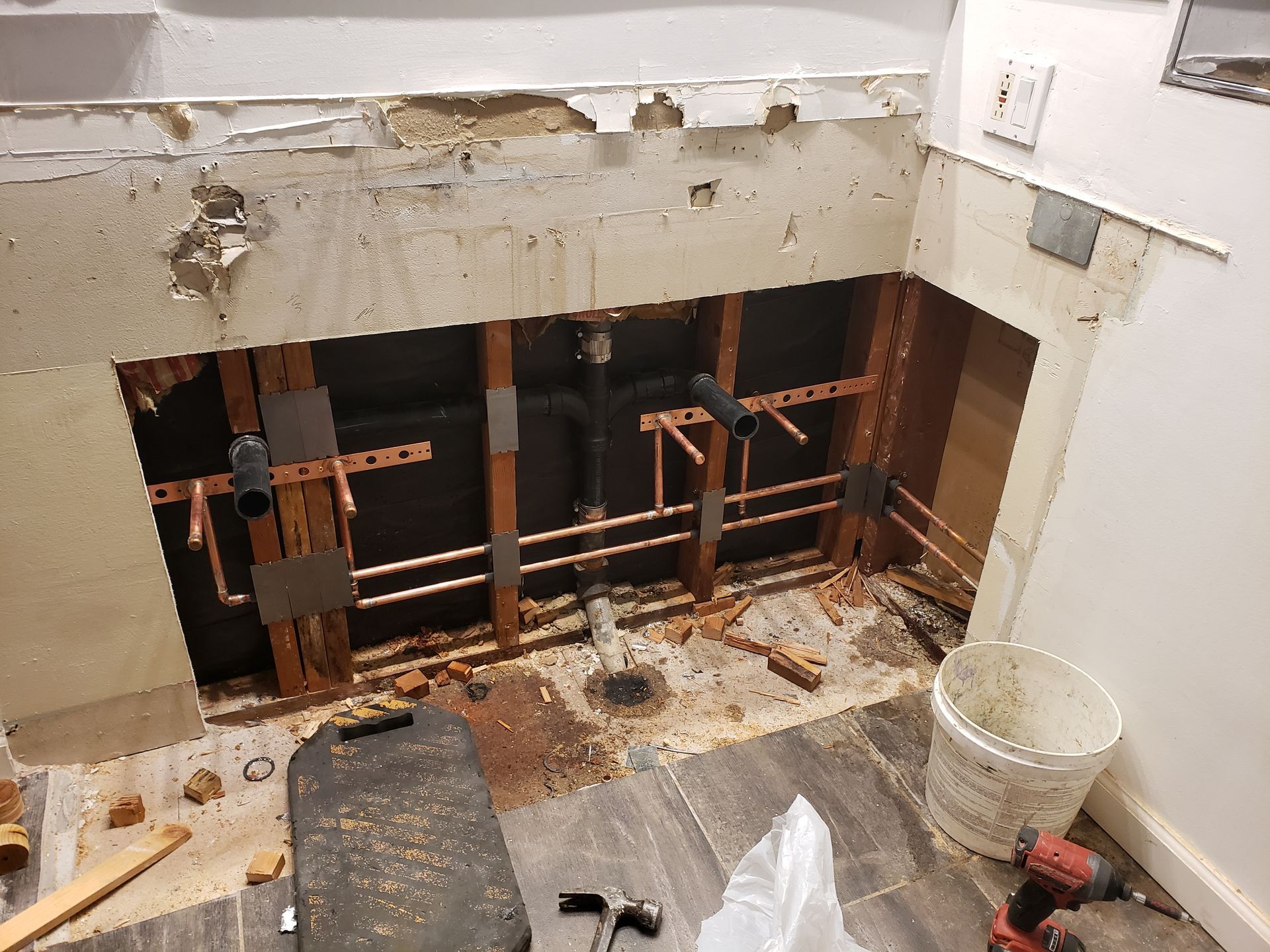 Bathroom wall opened to exposed plumbing and studs during renovation, with debris and tools on the floor