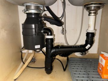 Plumber in blue uniform repairing a sink drain under a cabinet with a bucket below