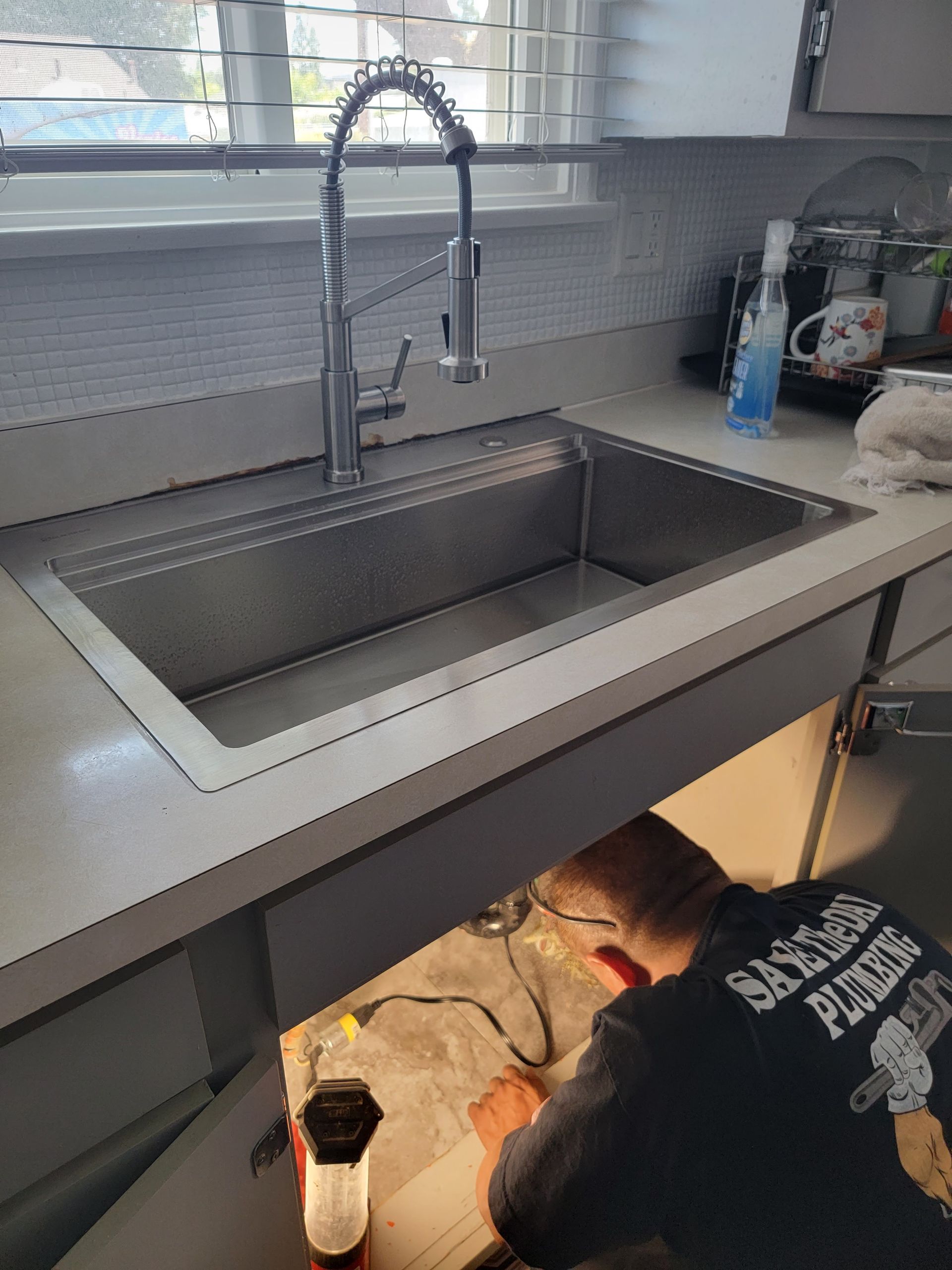 Person working under a kitchen sink with tools, next to a stainless steel basin and faucet.