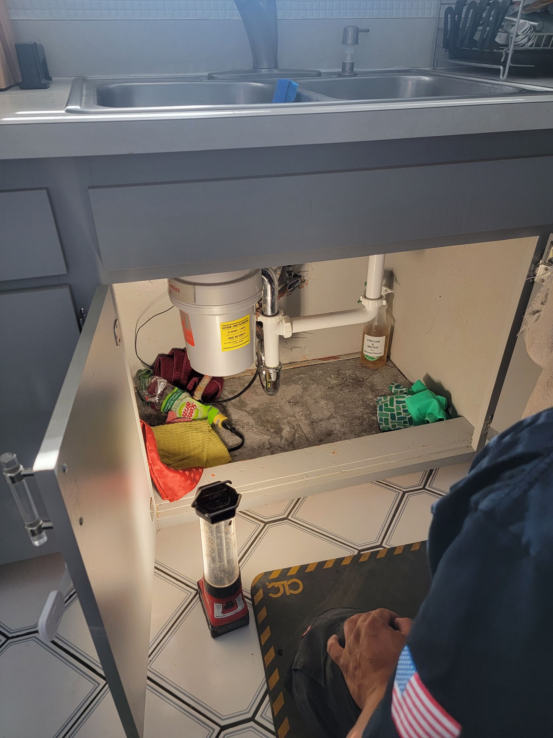 Under-sink cabinet with plumbing, cleaning supplies, and a person’s foot visible on the floor.