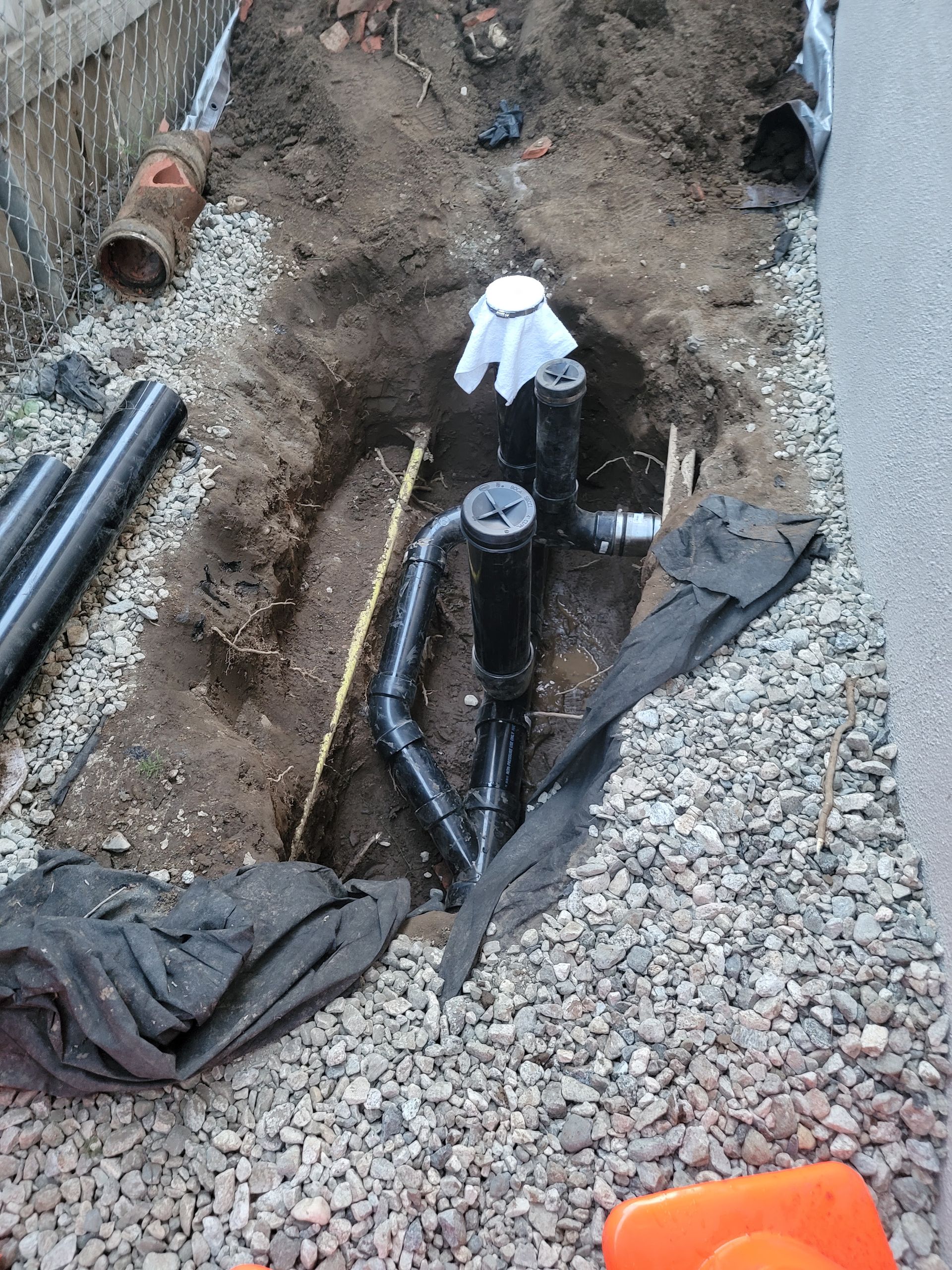 Trenched ground with exposed pipes and a black utility valve beside gravel and a small orange marker.