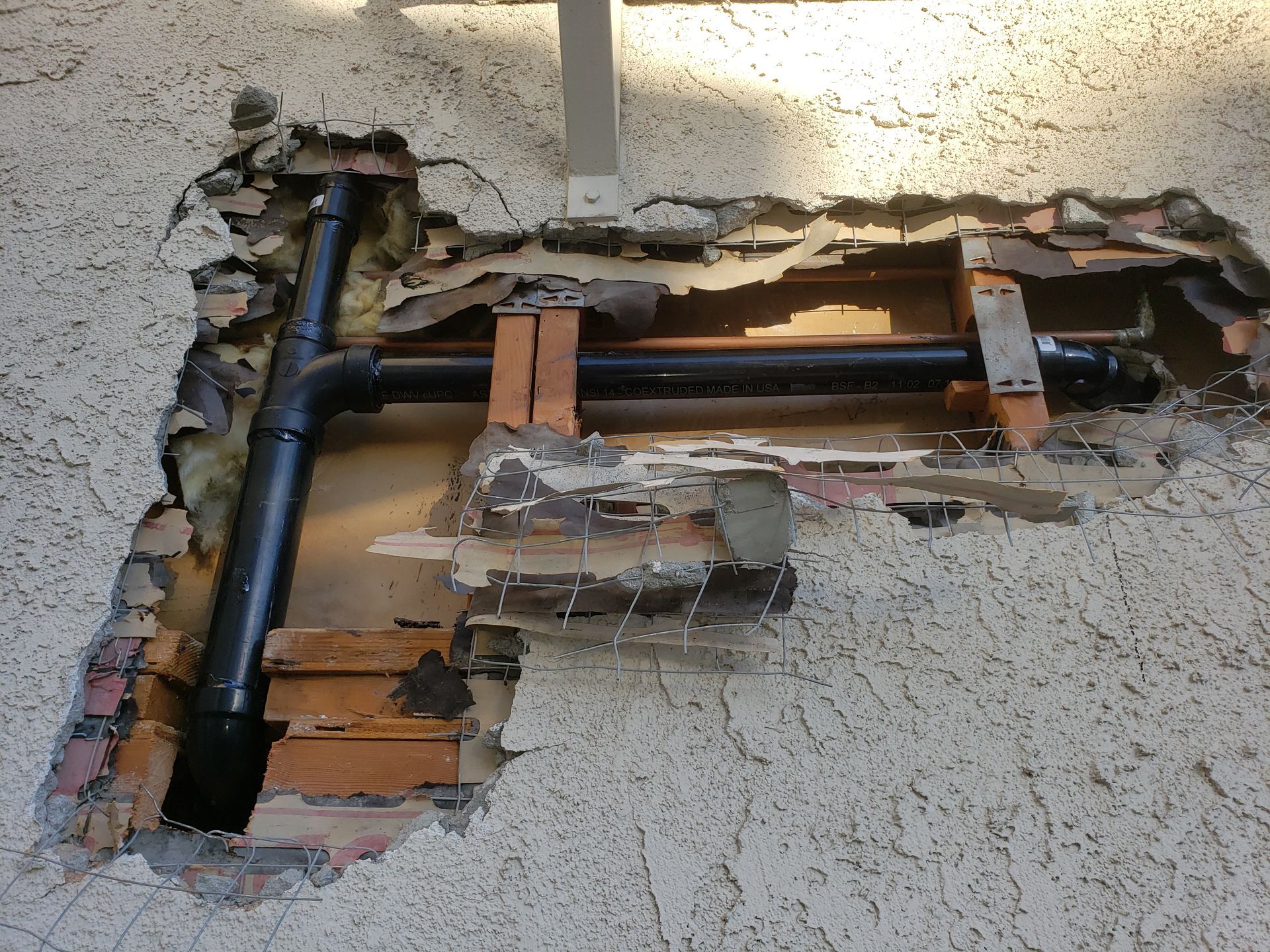 Exposed plumbing pipes inside a damaged wall with plaster and wood broken away