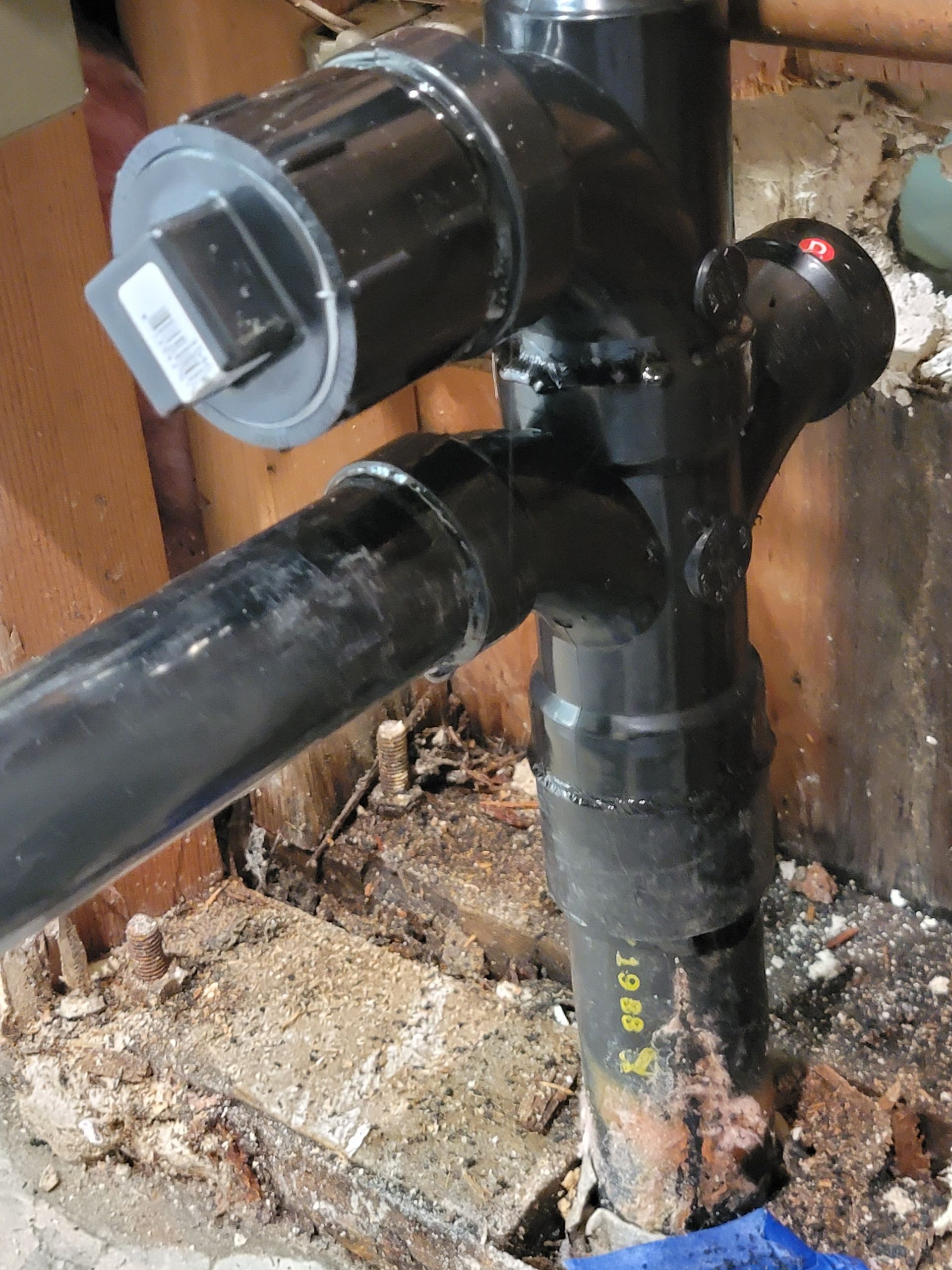 Black plastic plumbing pipes and a valve under a sink or floor opening, with a gray cap on top.