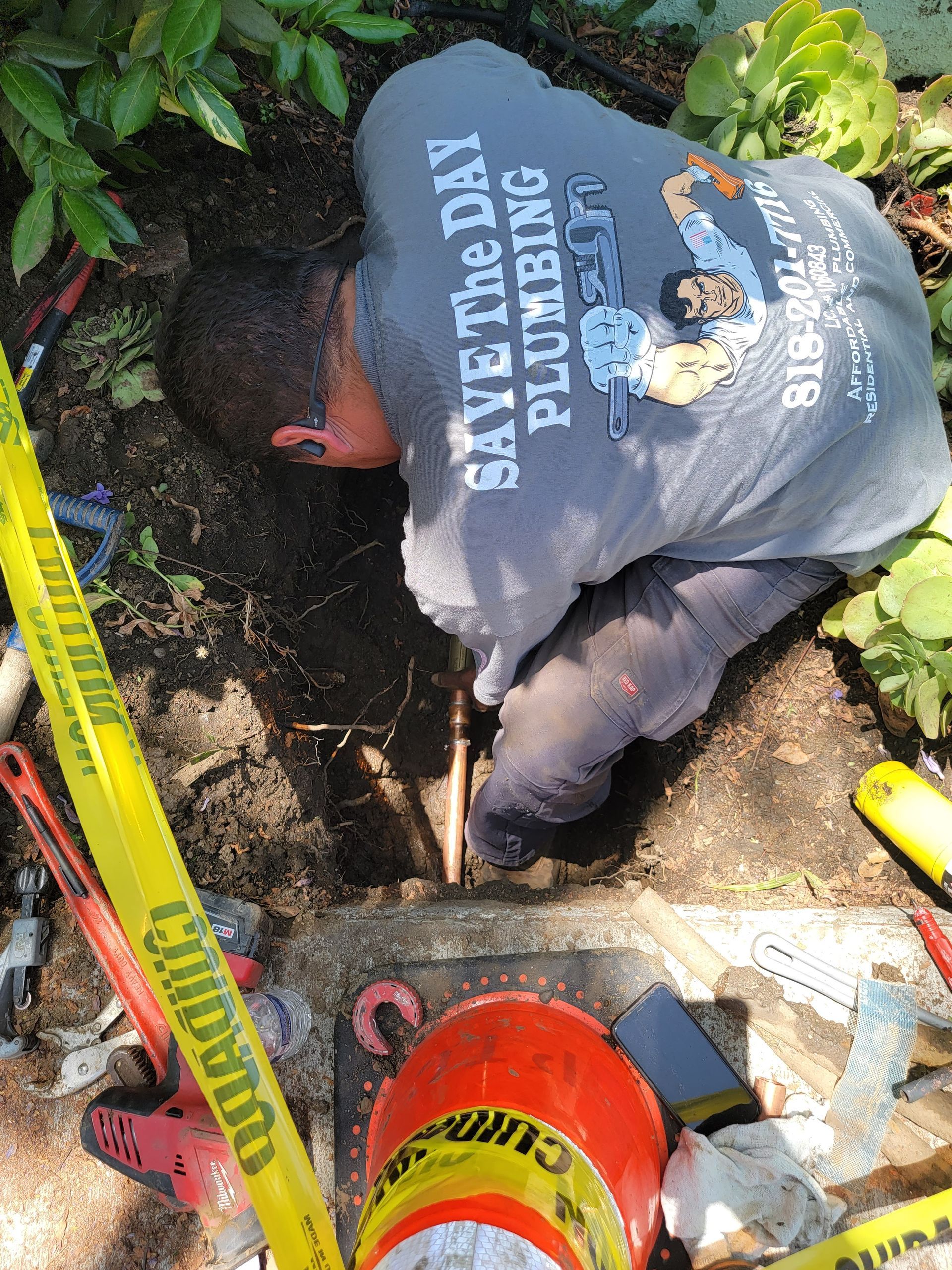 Worker in a trench beside a yellow tape and orange bucket, clearing mud with tools.