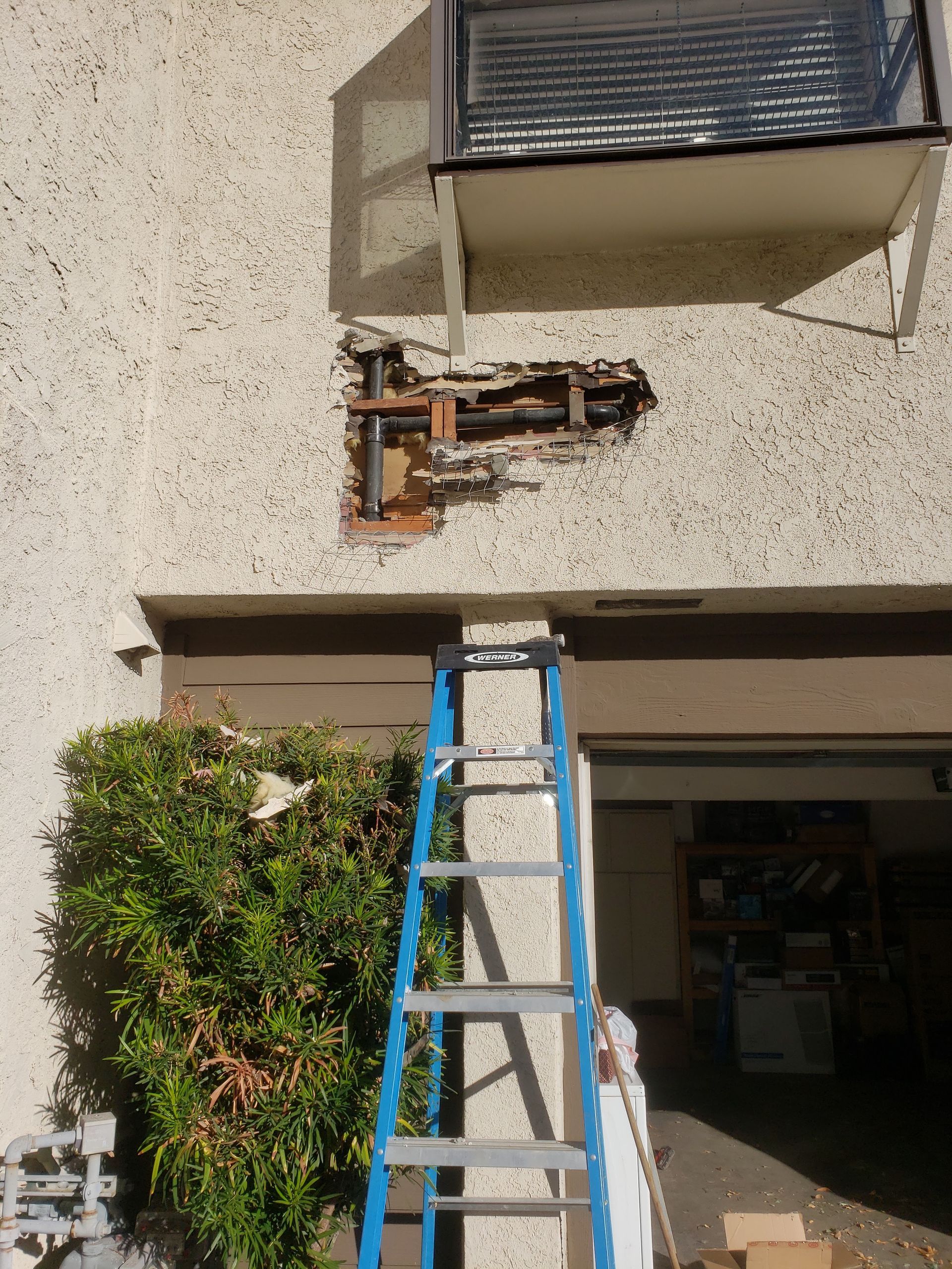 Blue ladder leaning against a stucco house beside a garage and balcony, with exposed wiring on the wall