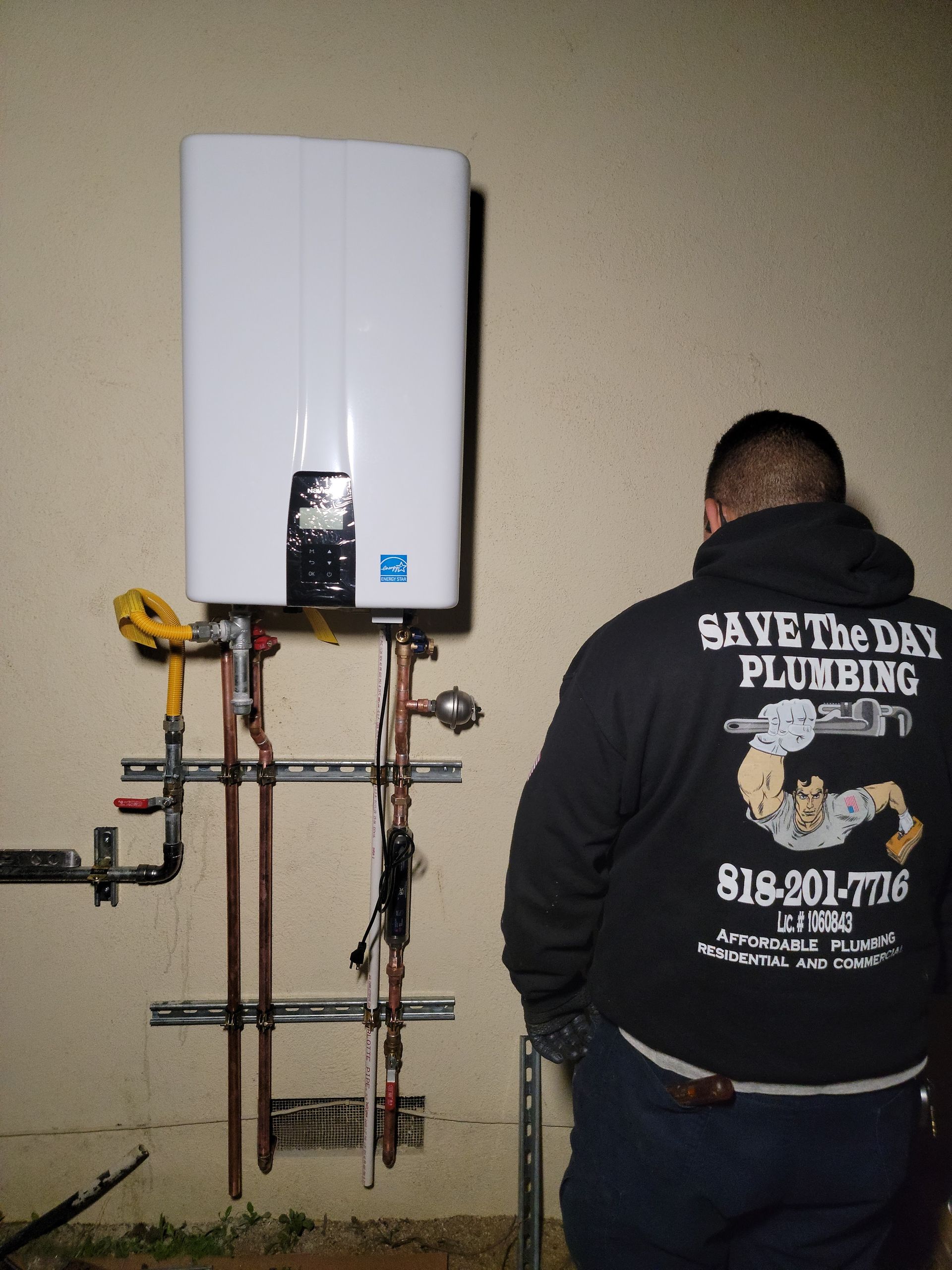 Plumber inspecting wall-mounted water heater and exposed pipes; man in black jacket stands beside the installation.