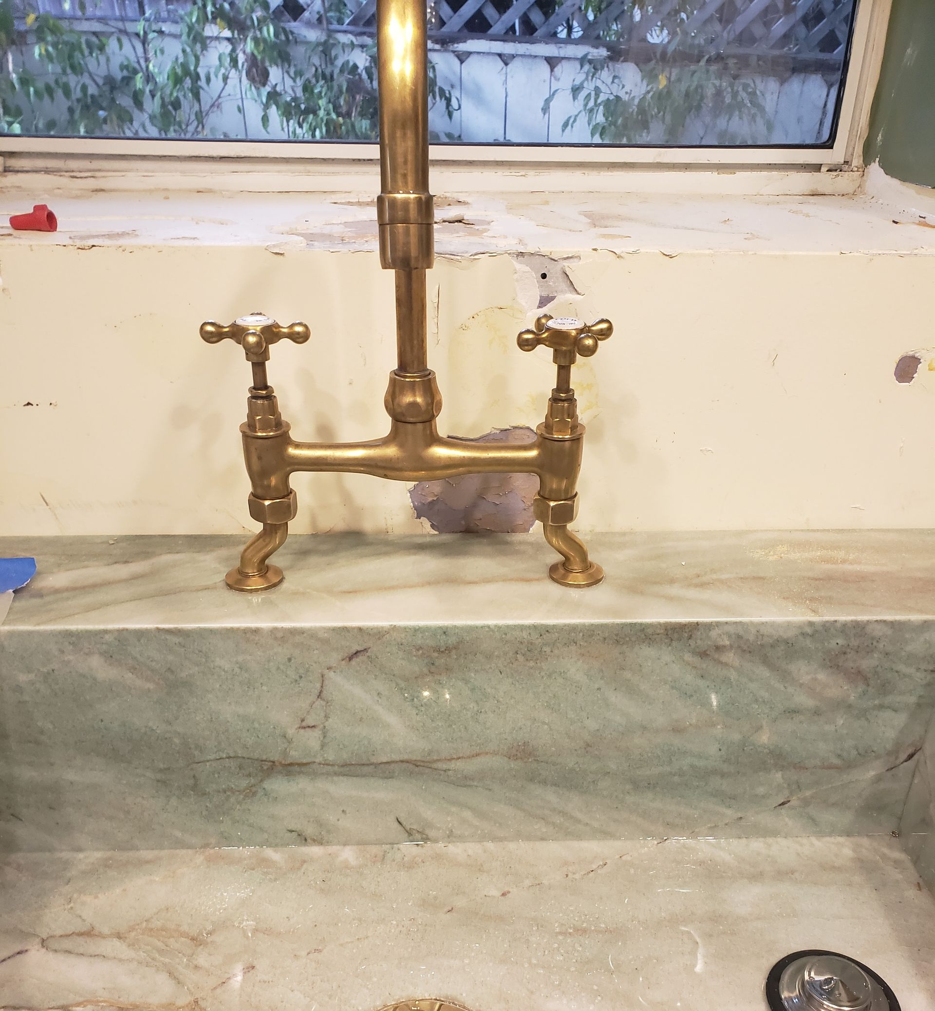 Old brass faucet and sink beneath a window in a worn kitchen area
