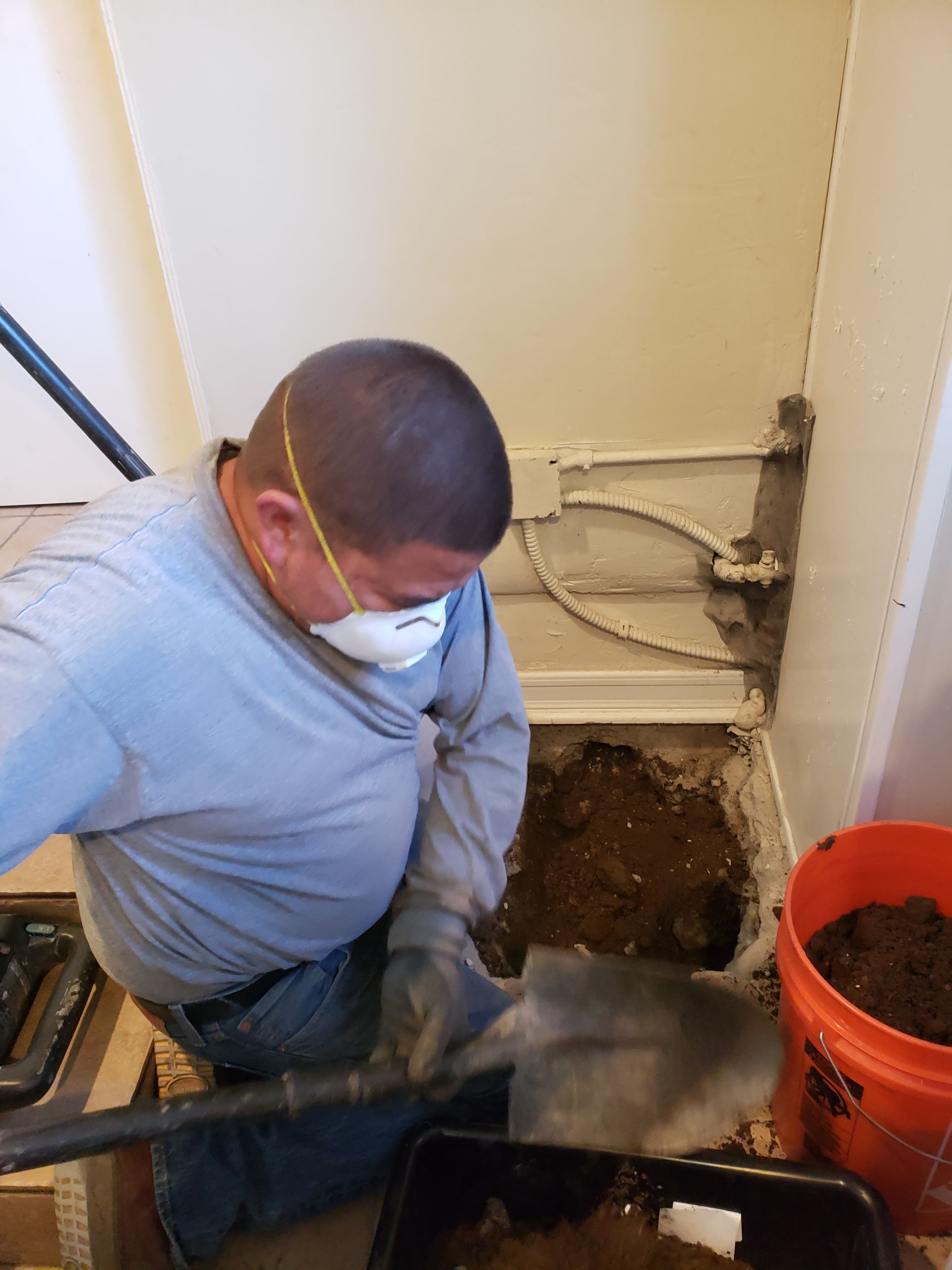 Worker using a shovel to dig in a small utility closet next to an orange bucket.