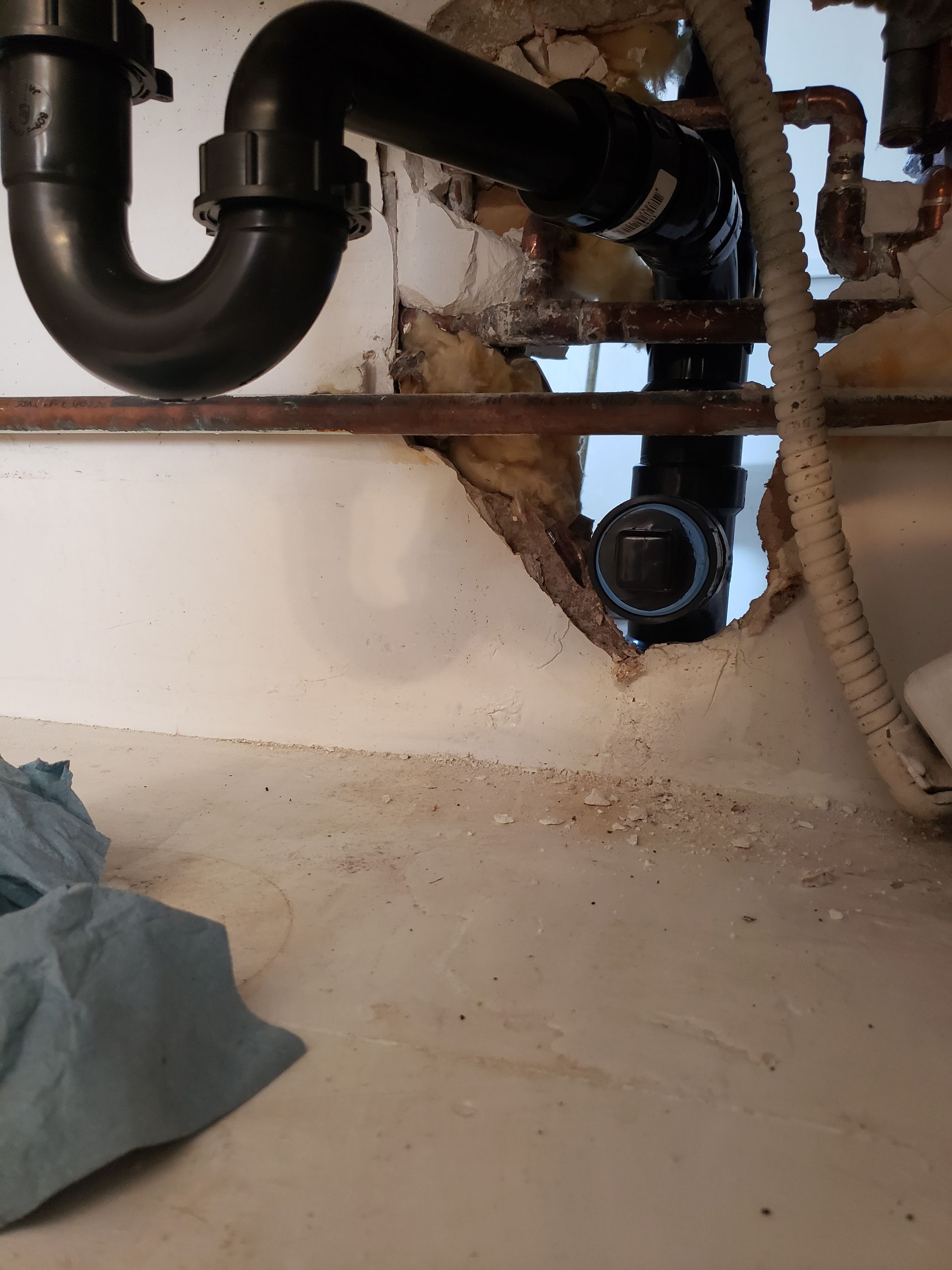 Under-sink plumbing with a black drain pipe, water damage, and debris on the floor