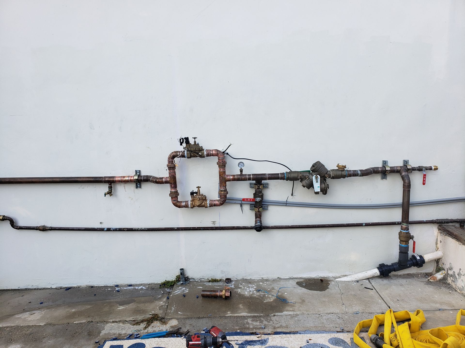 Wall-mounted pipes and valves on a white exterior wall, with rust, debris, and a yellow hose reel below.