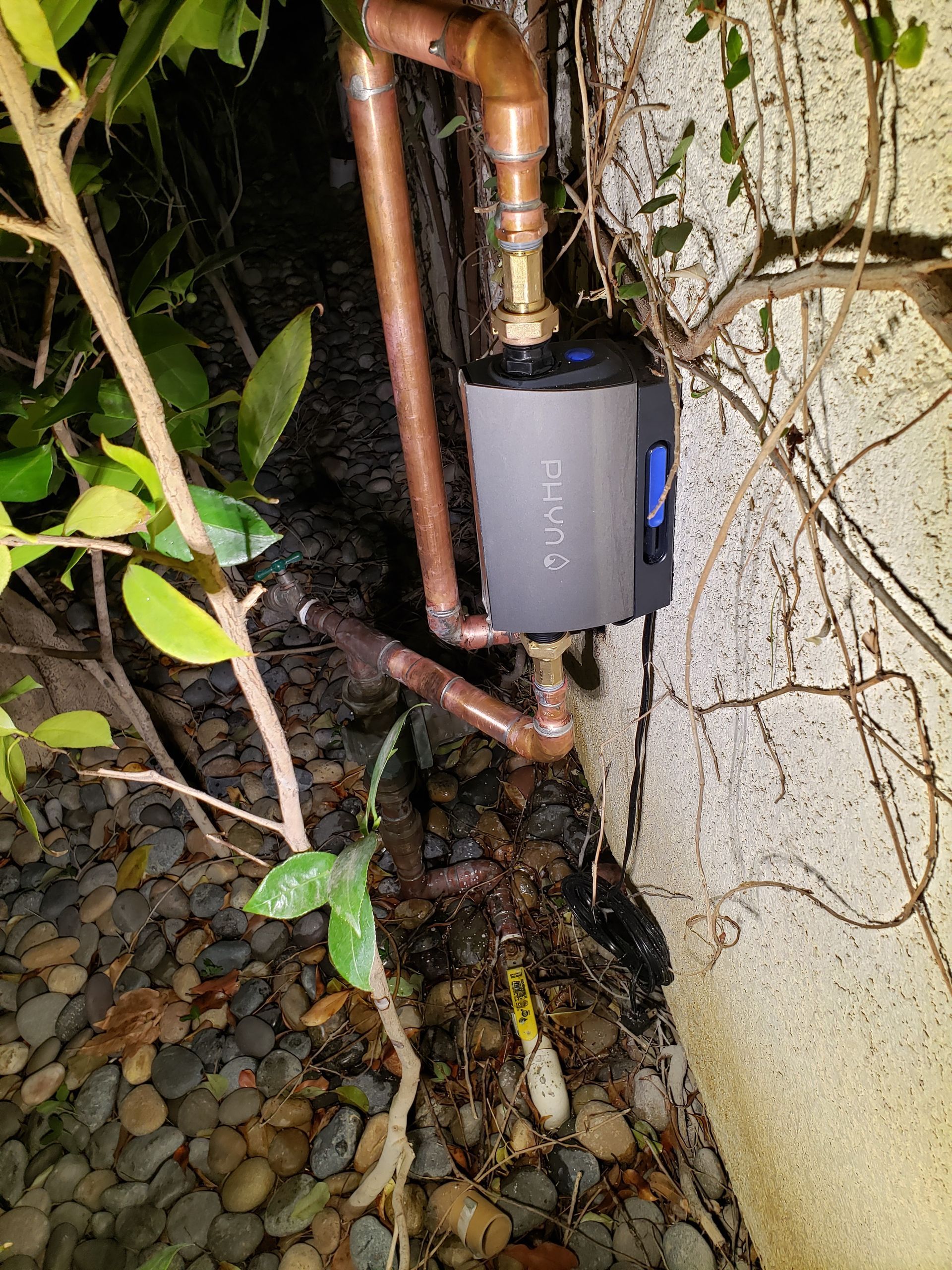 Outdoor electrical device mounted beside a wall, with copper pipes and vines over gravel.