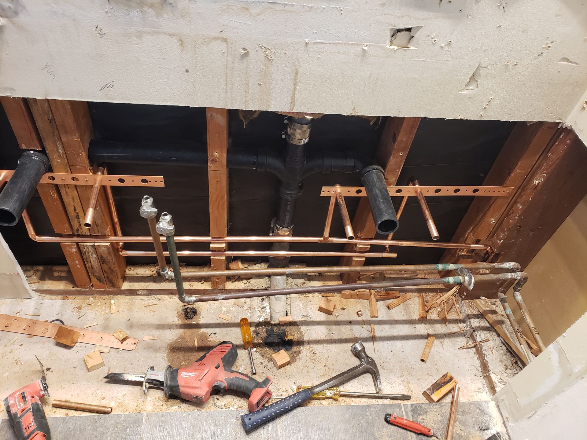Bathroom wall opened for plumbing repair, with exposed pipes, studs, tools, and debris on the floor.
