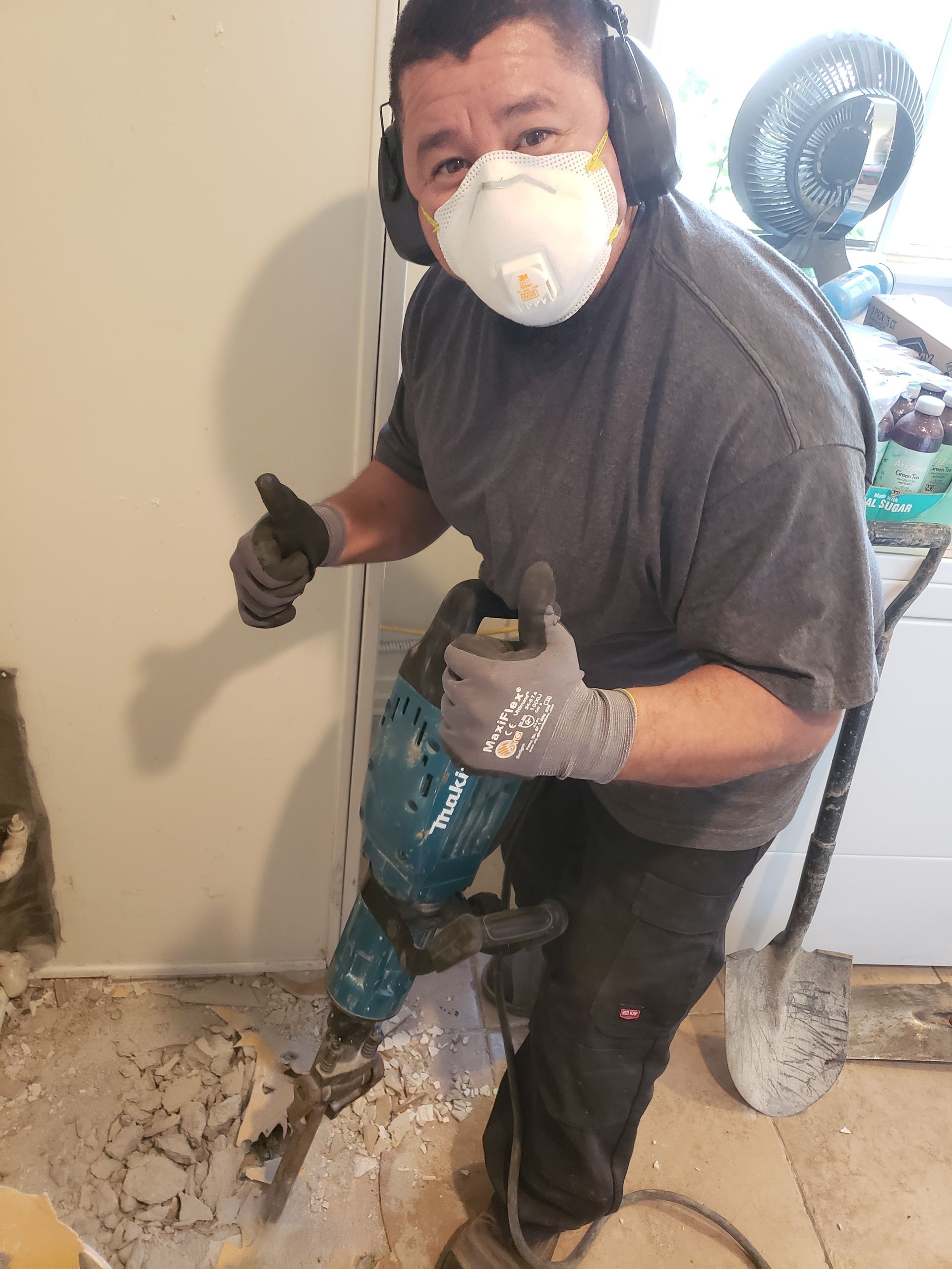 Worker using a jackhammer to break concrete indoors, wearing a respirator, gloves, and earmuffs.