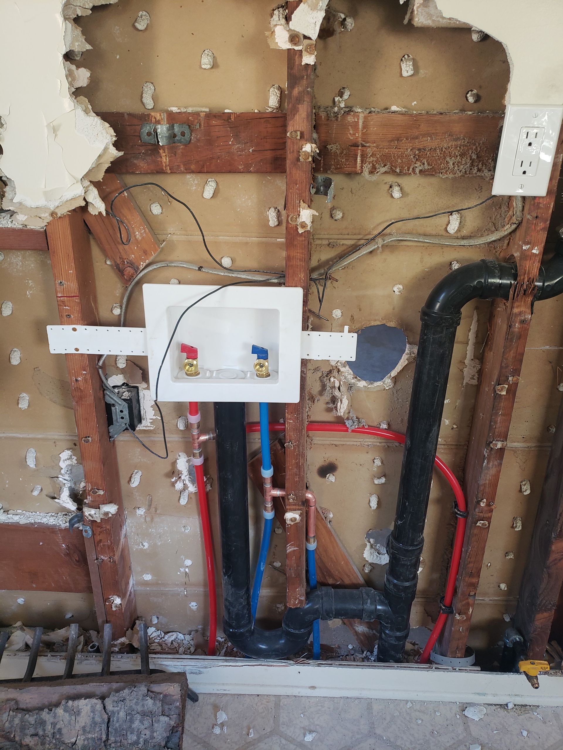 Open wall with exposed plumbing and a white utility box among red and blue pipes