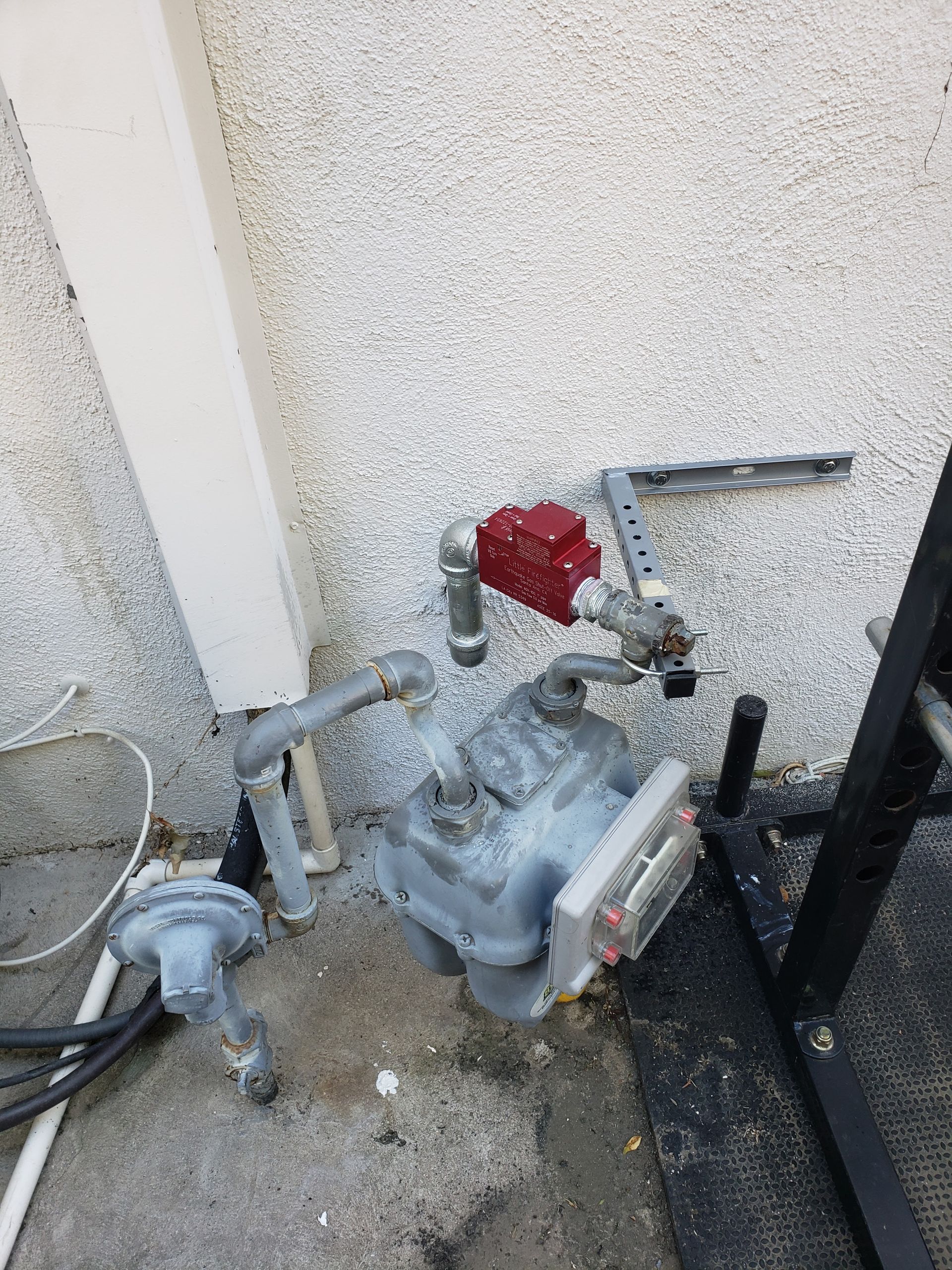 Outdoor gas meter with red valve and connected pipes beside a wall and fence.
