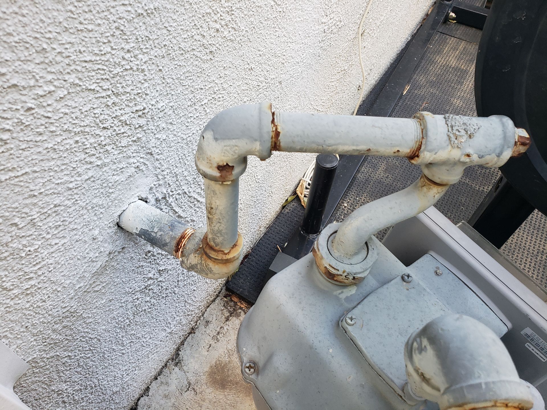 Rusty white pipes connected to a gray utility unit beside a stucco wall outdoors