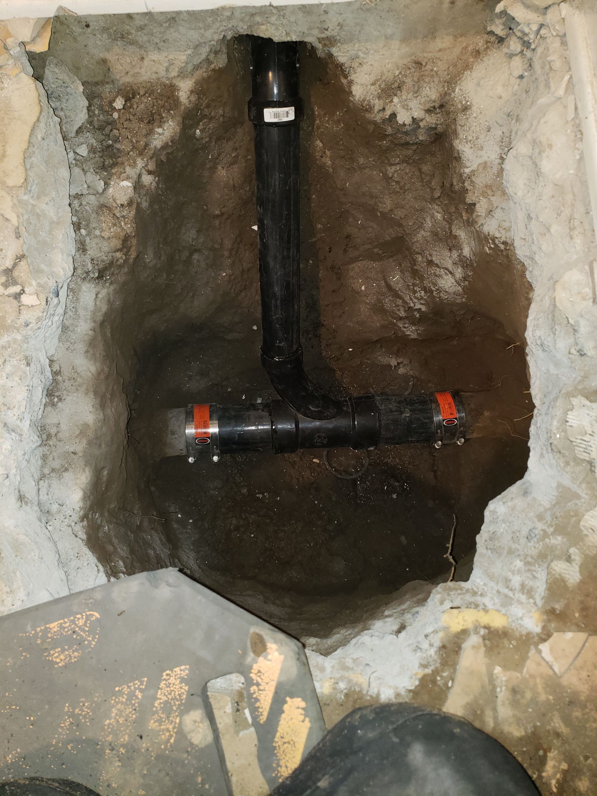 Excavated hole showing black PVC pipes with orange couplings in a concrete floor