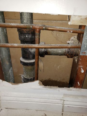 Rusty plumbing pipes inside a wall opening with broken white trim and exposed insulation.