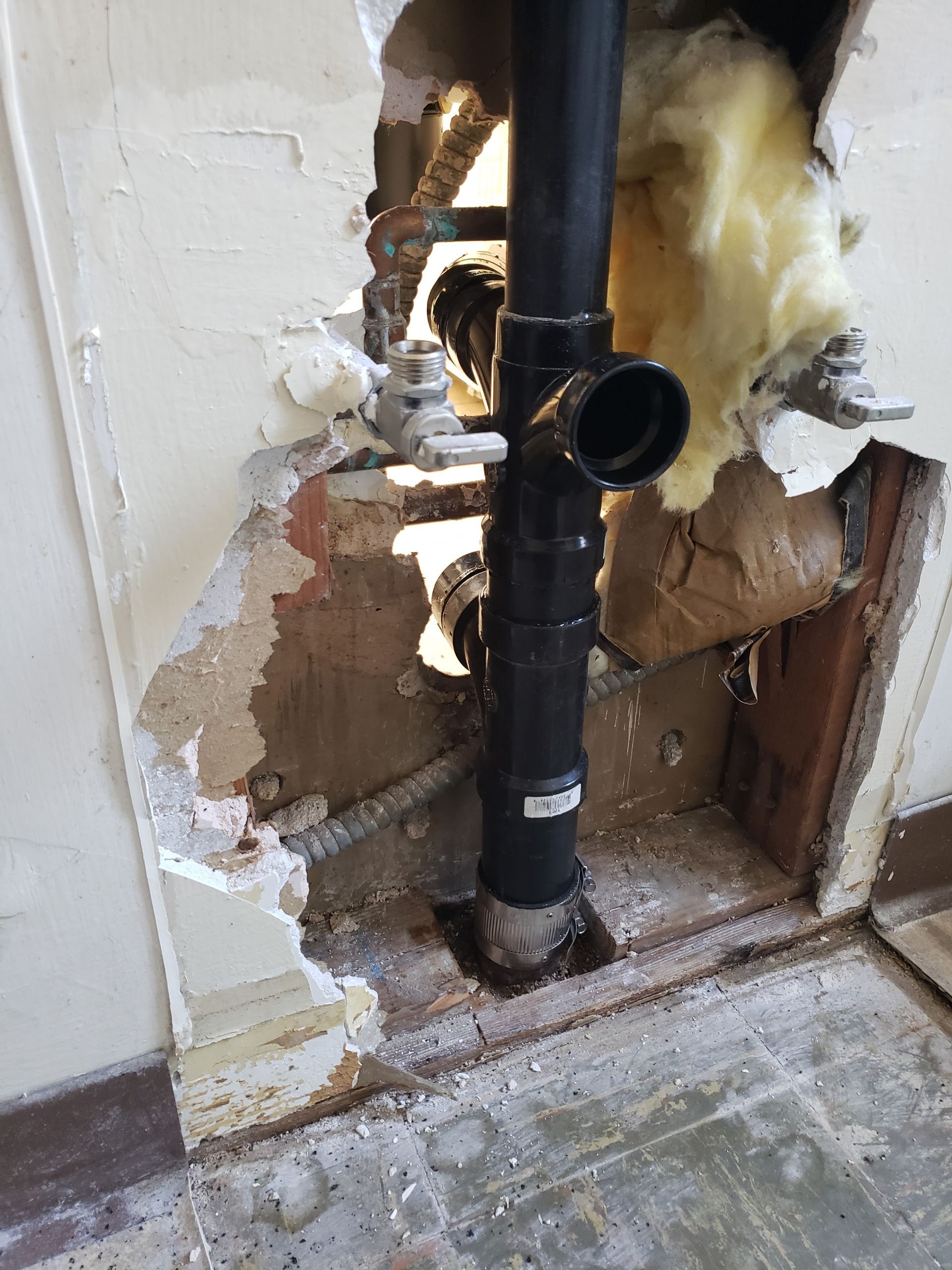 Damaged wall around a black pipe with exposed insulation and broken plaster near the floor