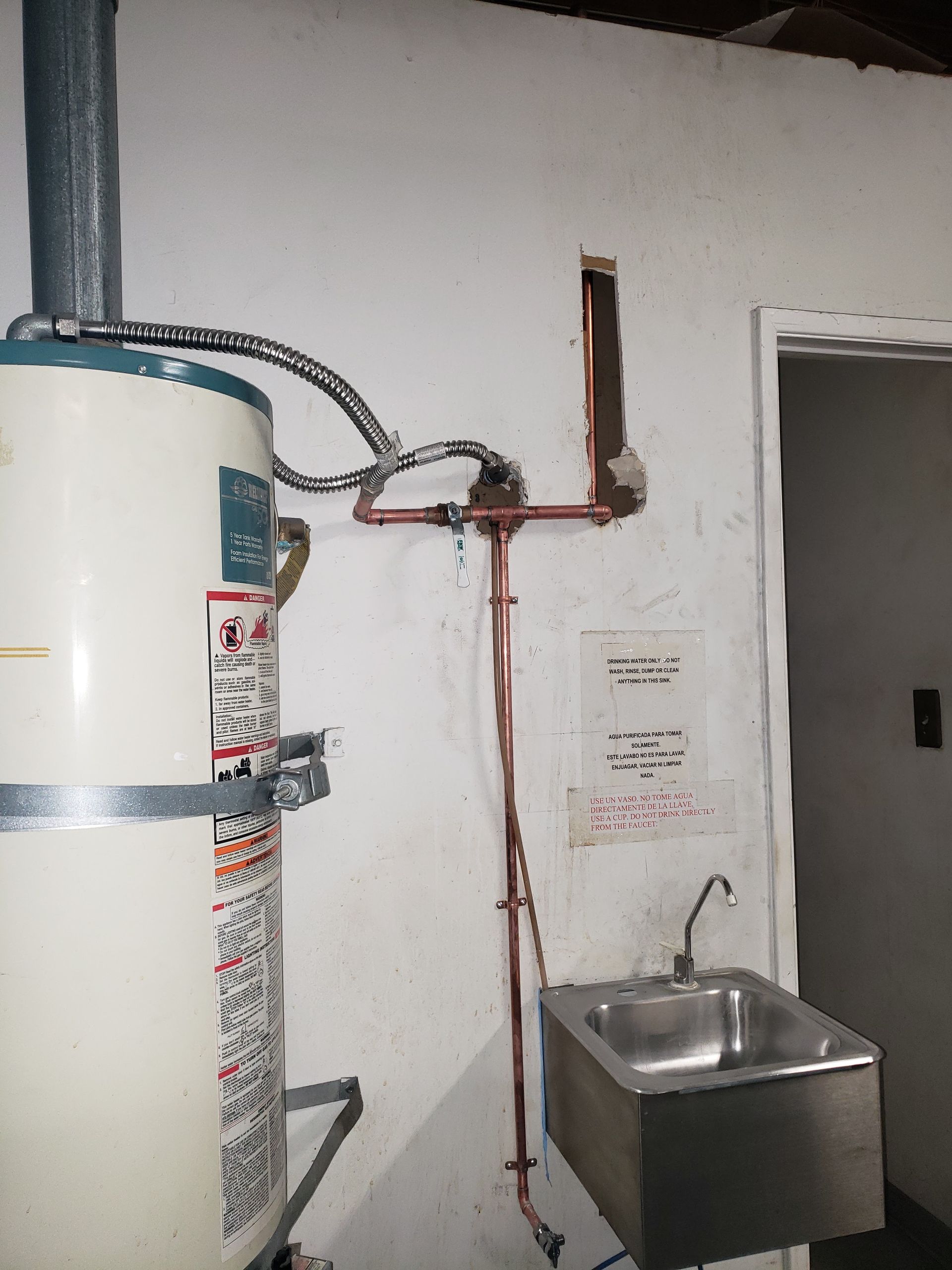 Water heater, exposed red pipes, and a small stainless-steel sink on a wall in a utility room