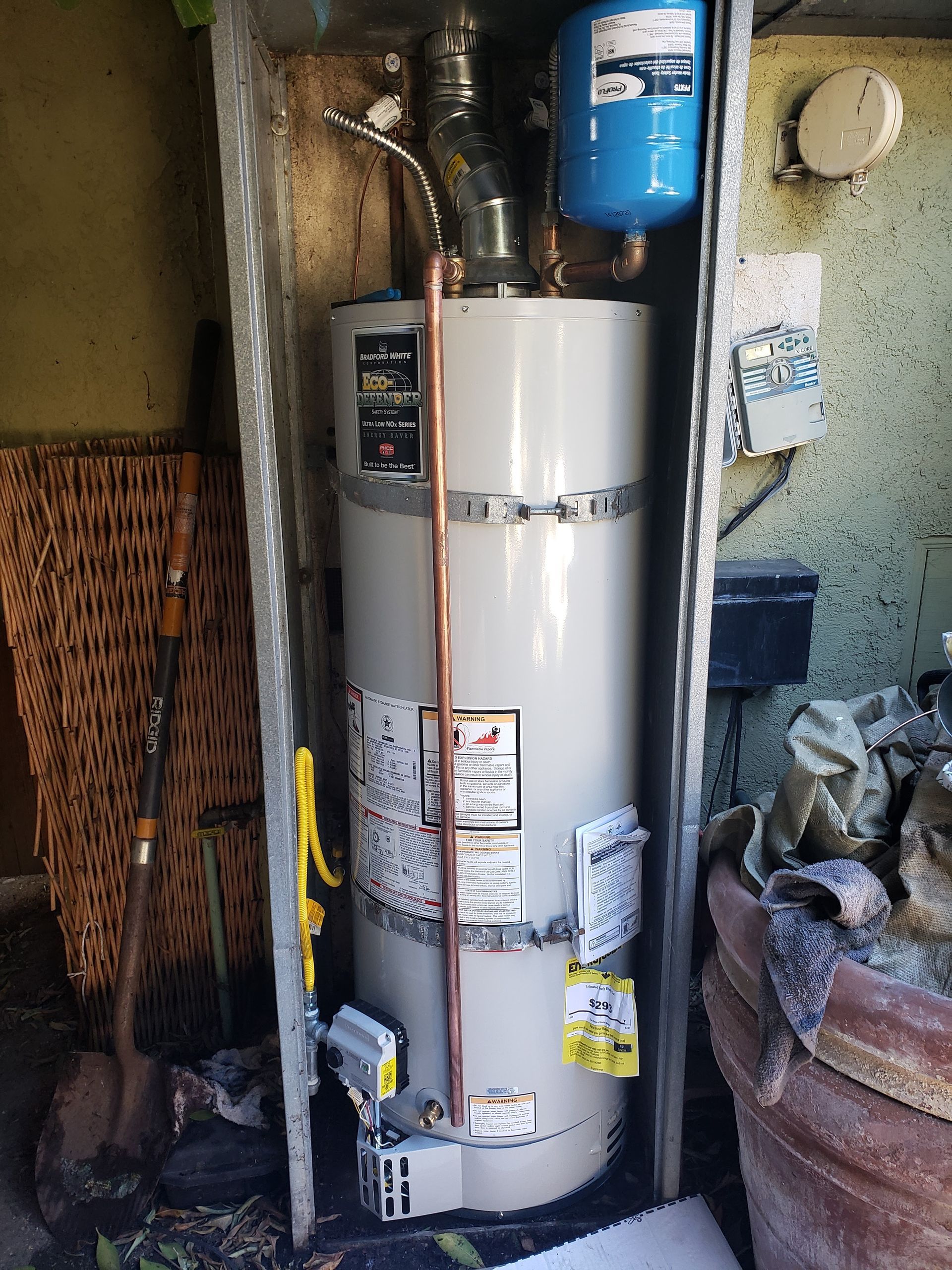 White water heater in a utility closet with pipes, a blue tank above, and a wicker basket beside it