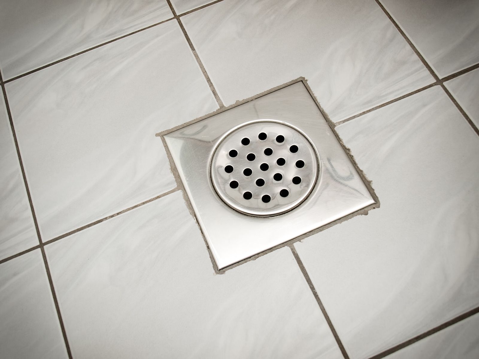 Bathroom floor drain with a round metal grate set in white tile.