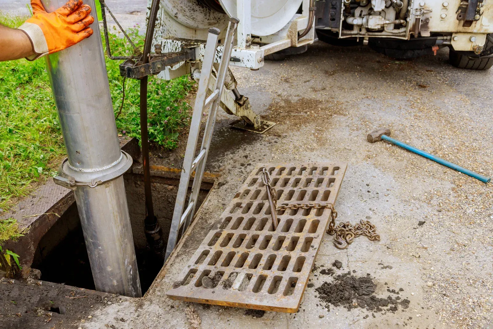 Worker opening a street drain beside a utility truck, with a metal grate and a long blue tool nearby