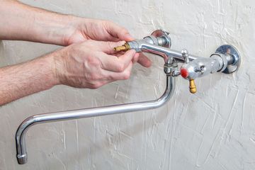 Hands adjusting a chrome wall-mounted faucet with a curved pipe against a textured white wall