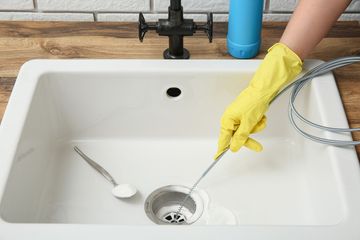 Gloved hand unclogs a kitchen sink drain with a cleaning tool in a white sink