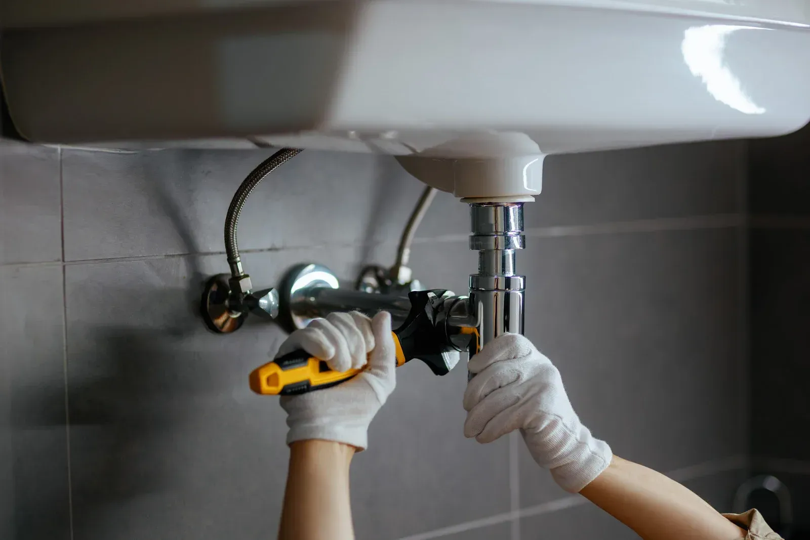 Hands repairing a sink drain under a white bathroom basin with a wrench and gloves