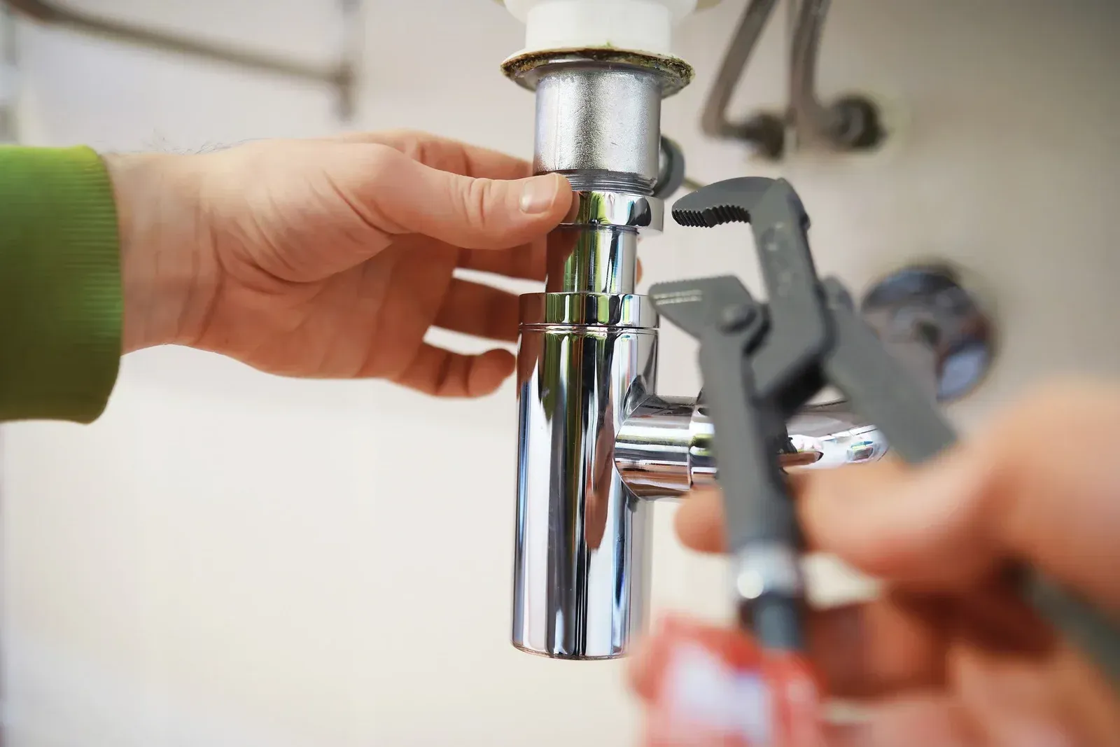 Hands tightening a chrome sink pipe with a wrench under a bathroom sink
