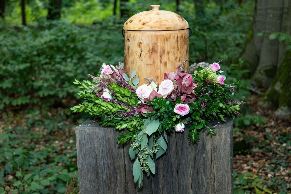 A natural birch wood cremation urn sits on a dark tree stump, surrounded by a ring of pink flowers and lush greenery.