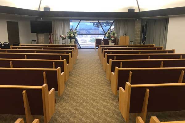 Empty rows of wooden pews face a central aisle leading to a stage with floral arrangements and a window view.
