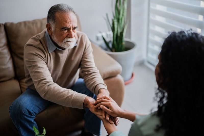 An older man listens as a woman holds his hand in comfort on a living room sofa