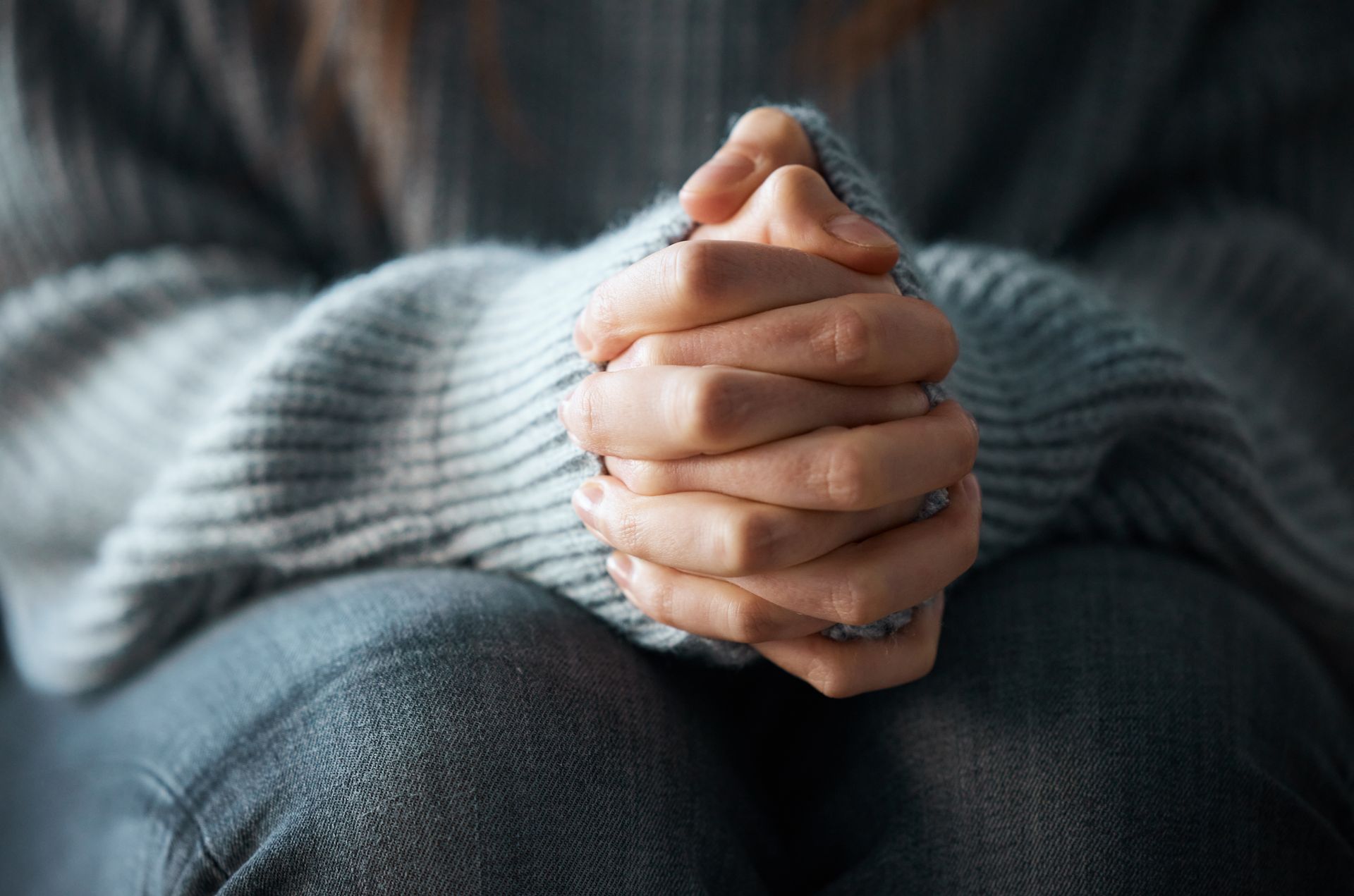 Clasped hands resting on a lap, wearing a gray sweater and dark jeans.