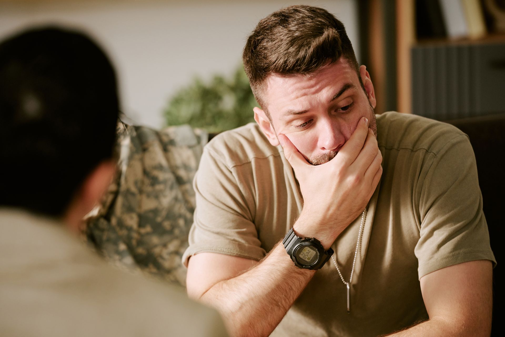 Man sitting with his hand over his mouth, looking distressed in a room with another person blurred in the foreground