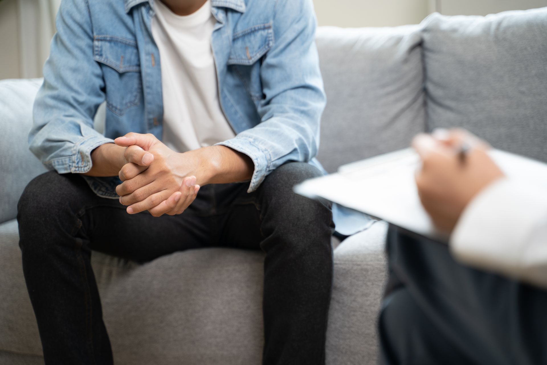 Person seated on a gray couch, hands clasped, in a counseling session with a clipboard in the foreground