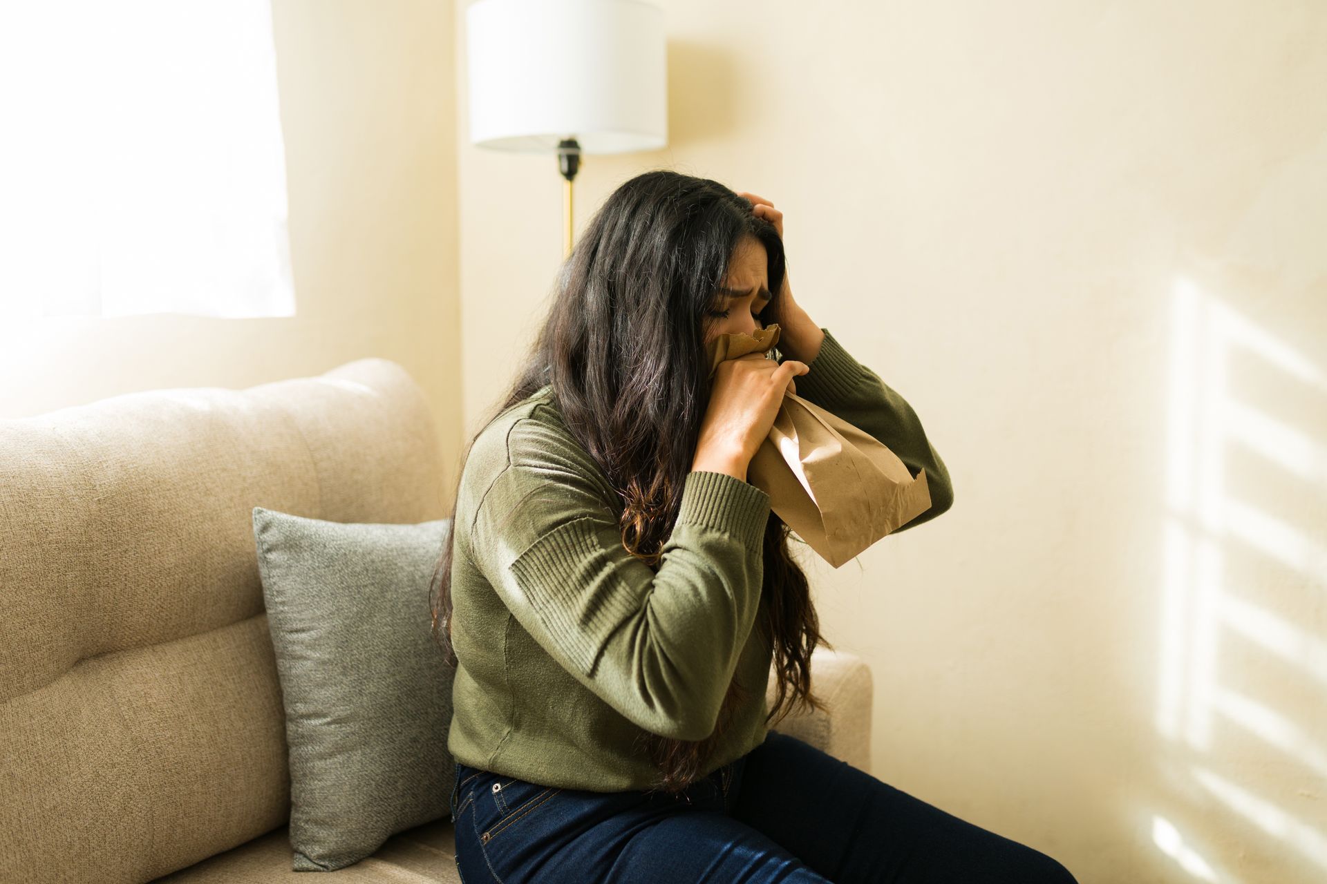 Person sitting on a couch, holding a paper bag to their face in a bright living room.