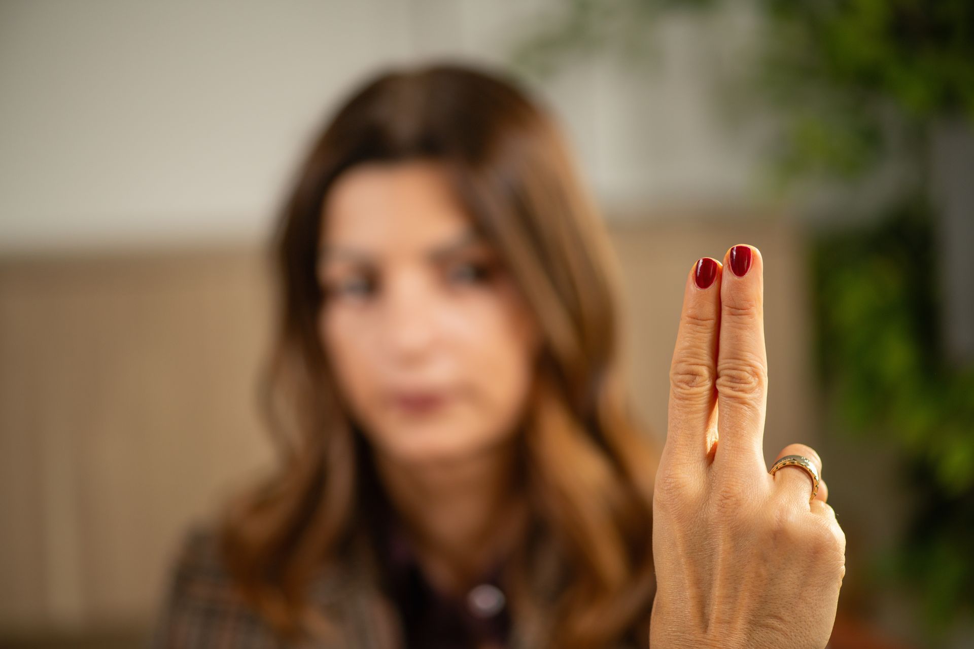 Two raised fingers with red nails in the foreground, blurred woman in the background indoors