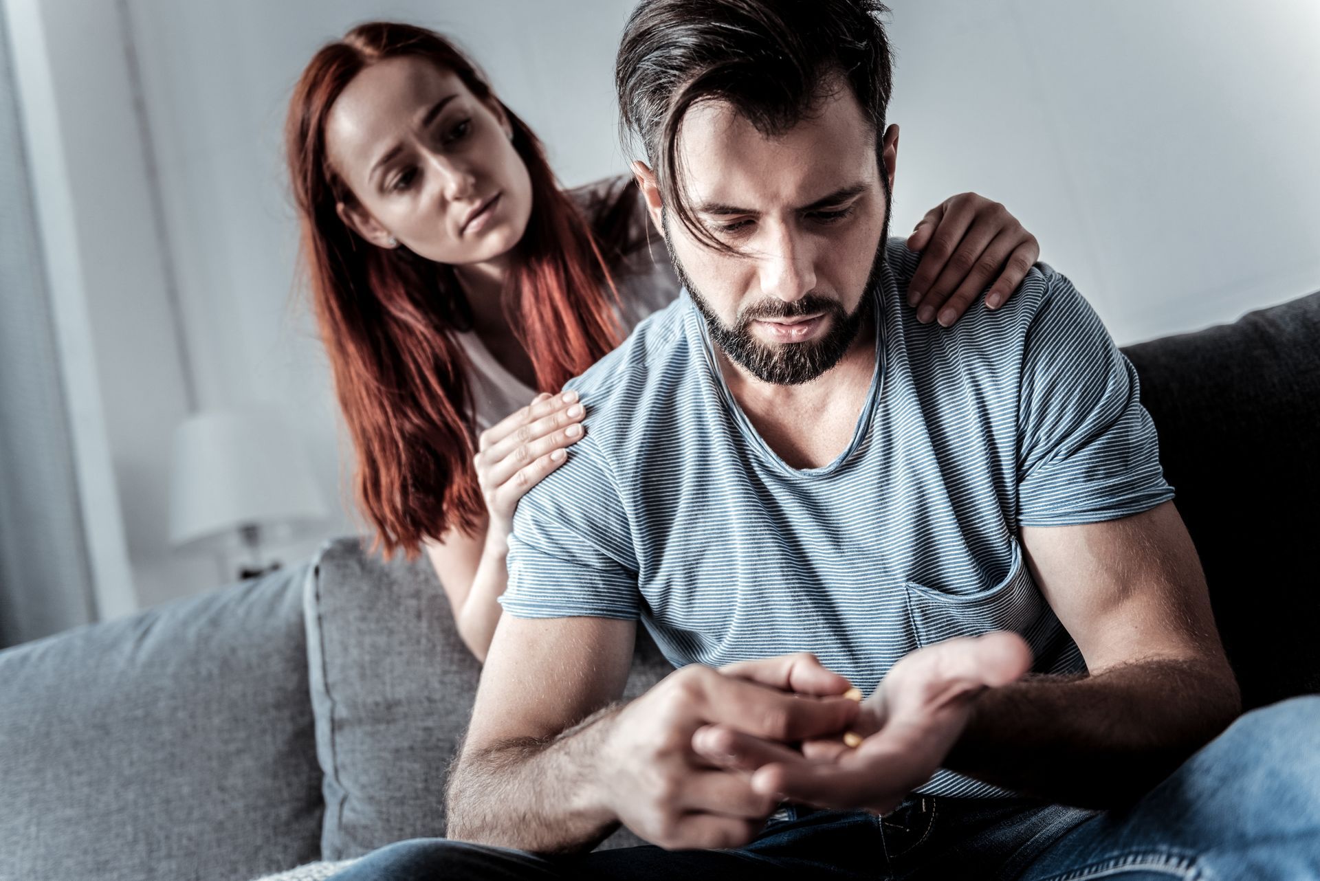 Two people sitting on a couch, one comforting the other while he looks down at his phone