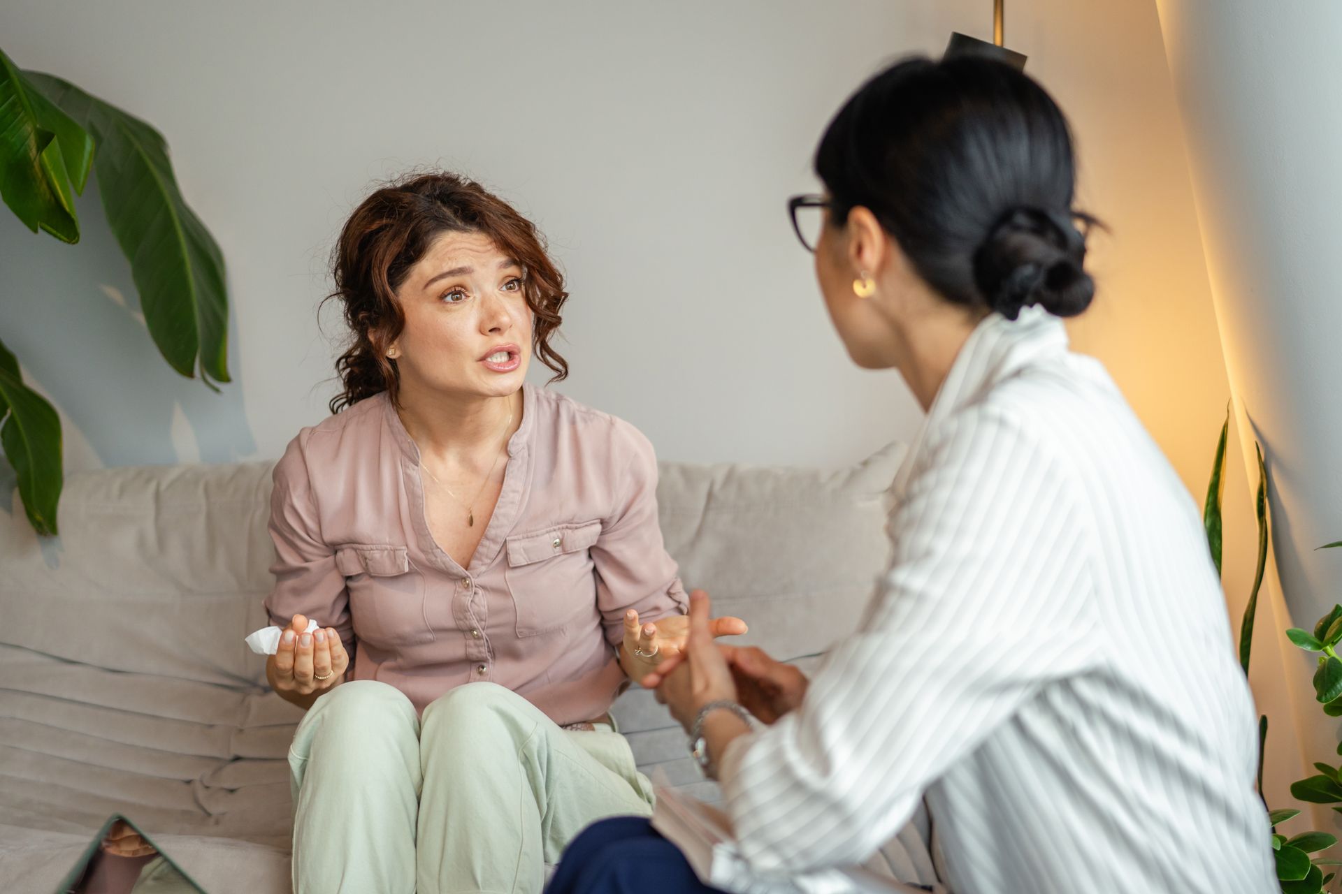 Two people talking on a couch in a bright living room, one holding a tissue while listening.