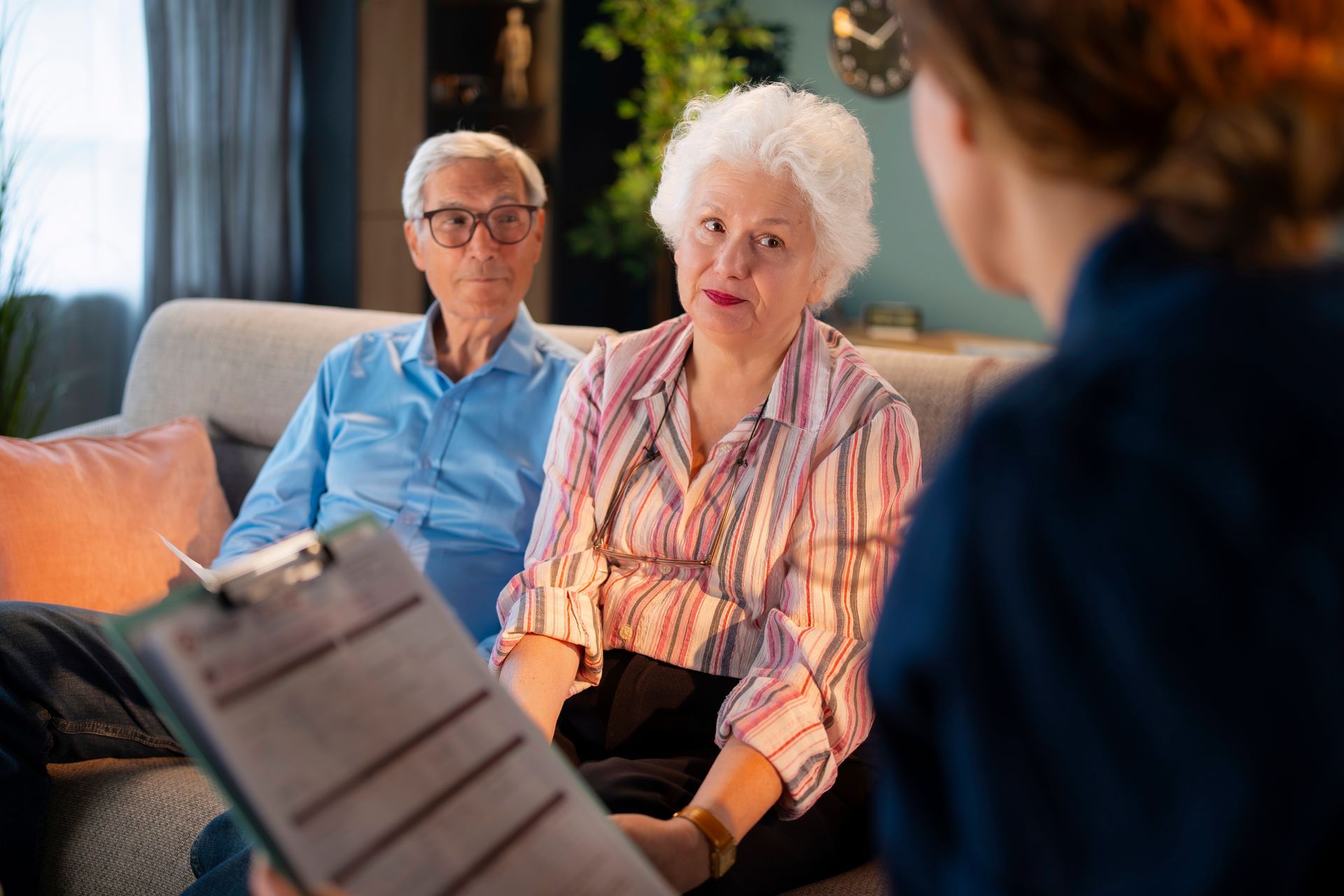 Elderly couple listening to a therapist in a living room, with a clipboard in the foreground.