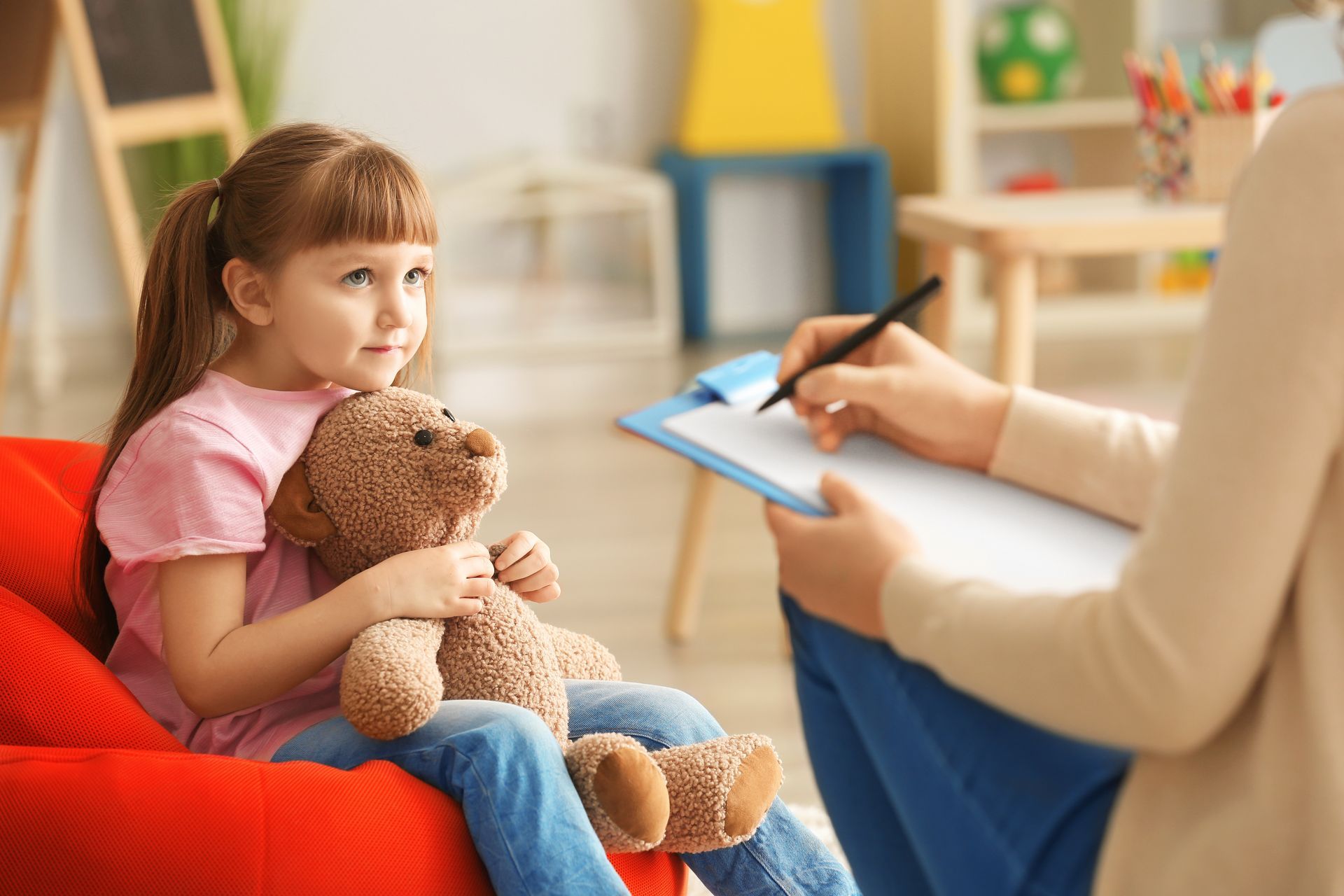 Child sitting in a red chair, holding a teddy bear during a counseling session