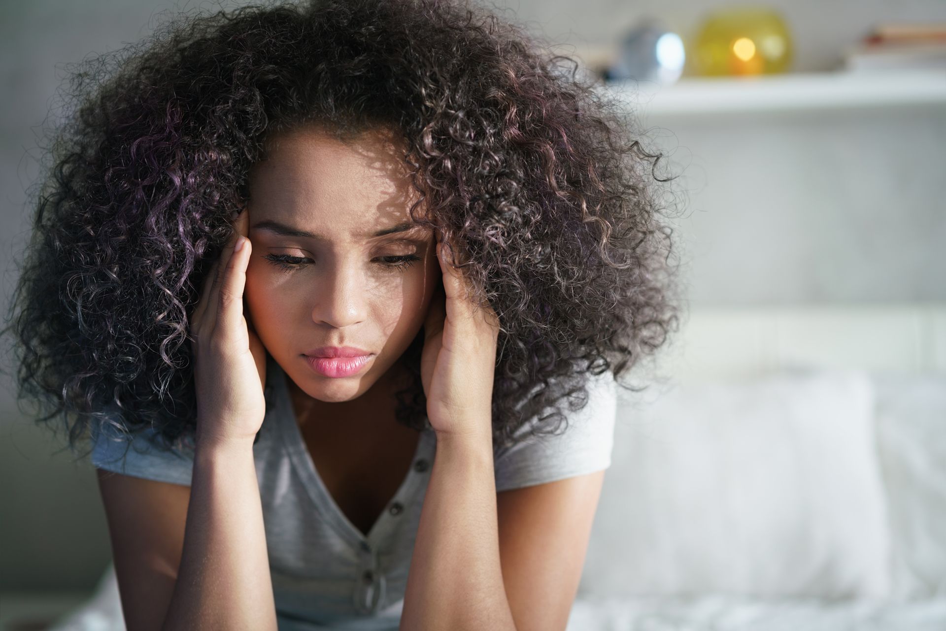 Woman with curly hair sitting on a bed, resting her head in her hands and looking down.