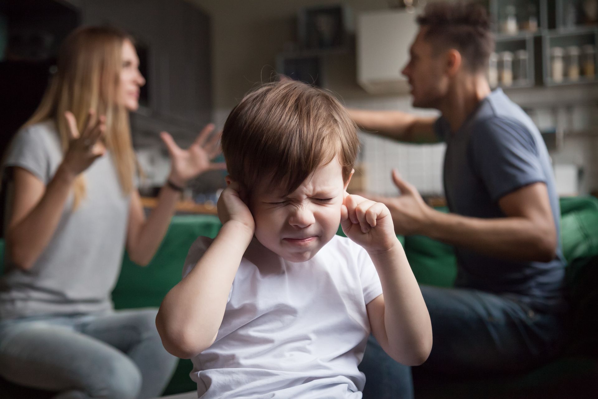 Child covering ears while parents argue in the background indoors